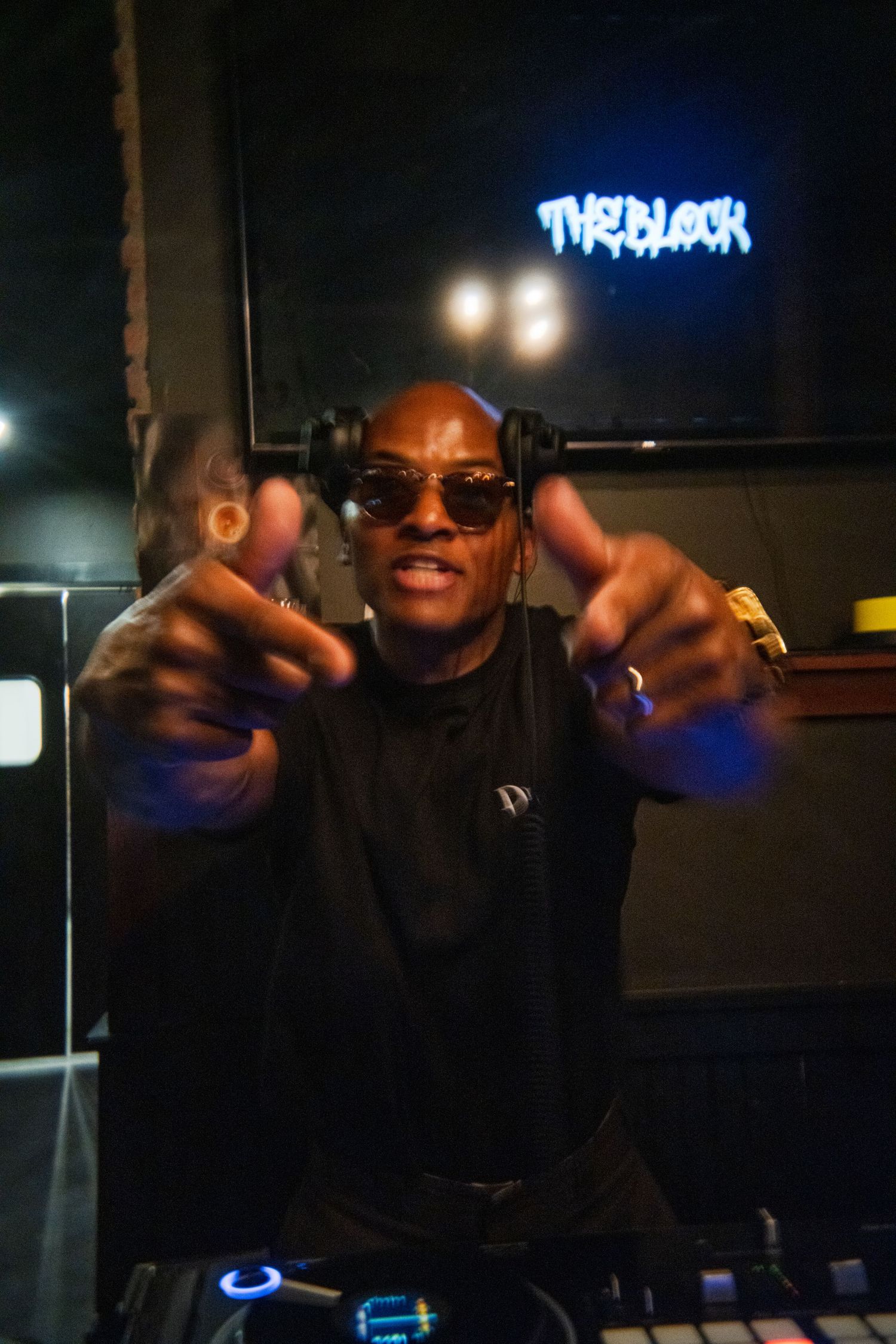 A DJ performs at a nightclub with neon signs and dark lighting visible in the background.