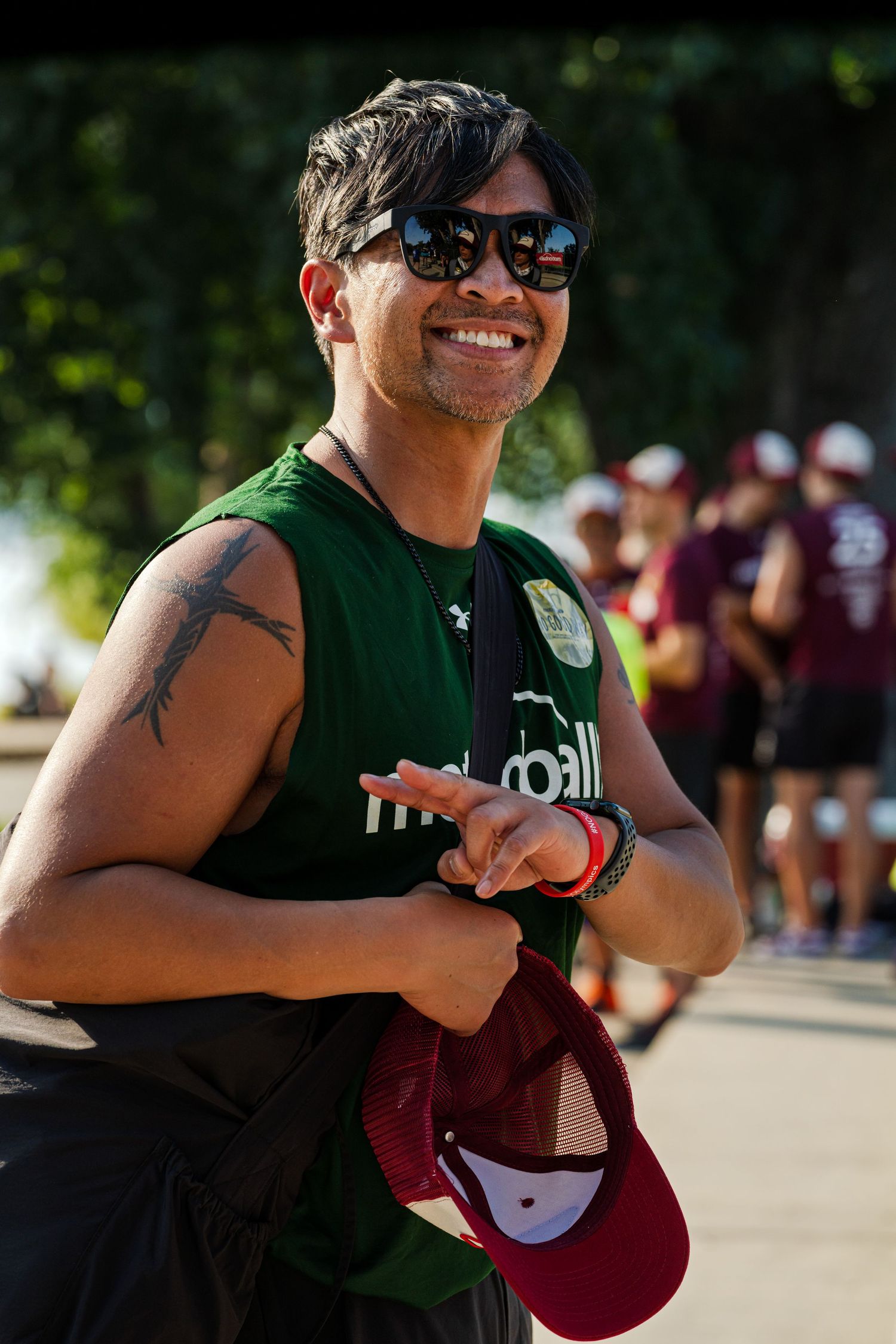 Smiling person in sunglasses and green sleeveless shirt holding a red cap at an outdoor event.