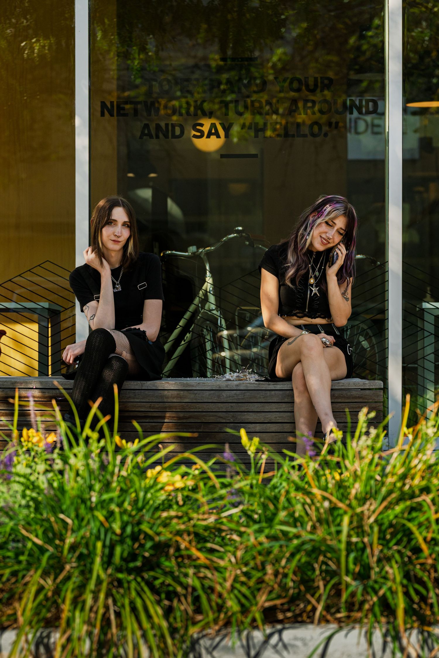 Two people sit on steps outside a storefront with greenery in the foreground during golden hour.