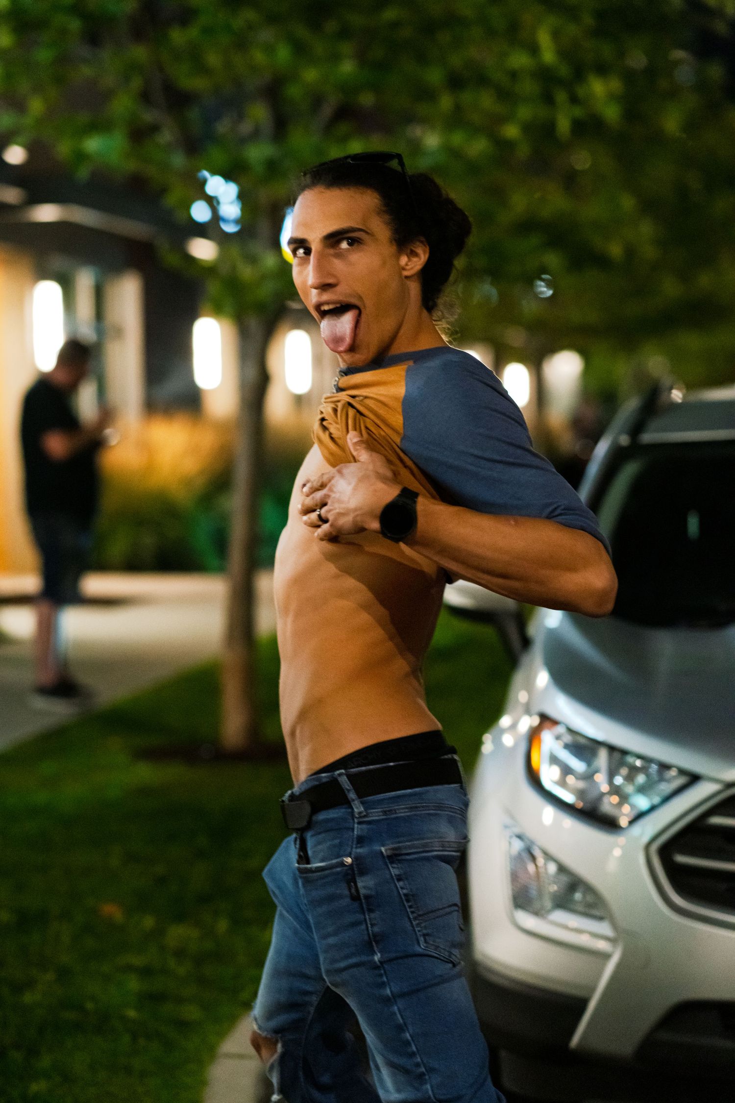 A playful nighttime portrait showing someone in jeans lifting their shirt in front of a parked car.