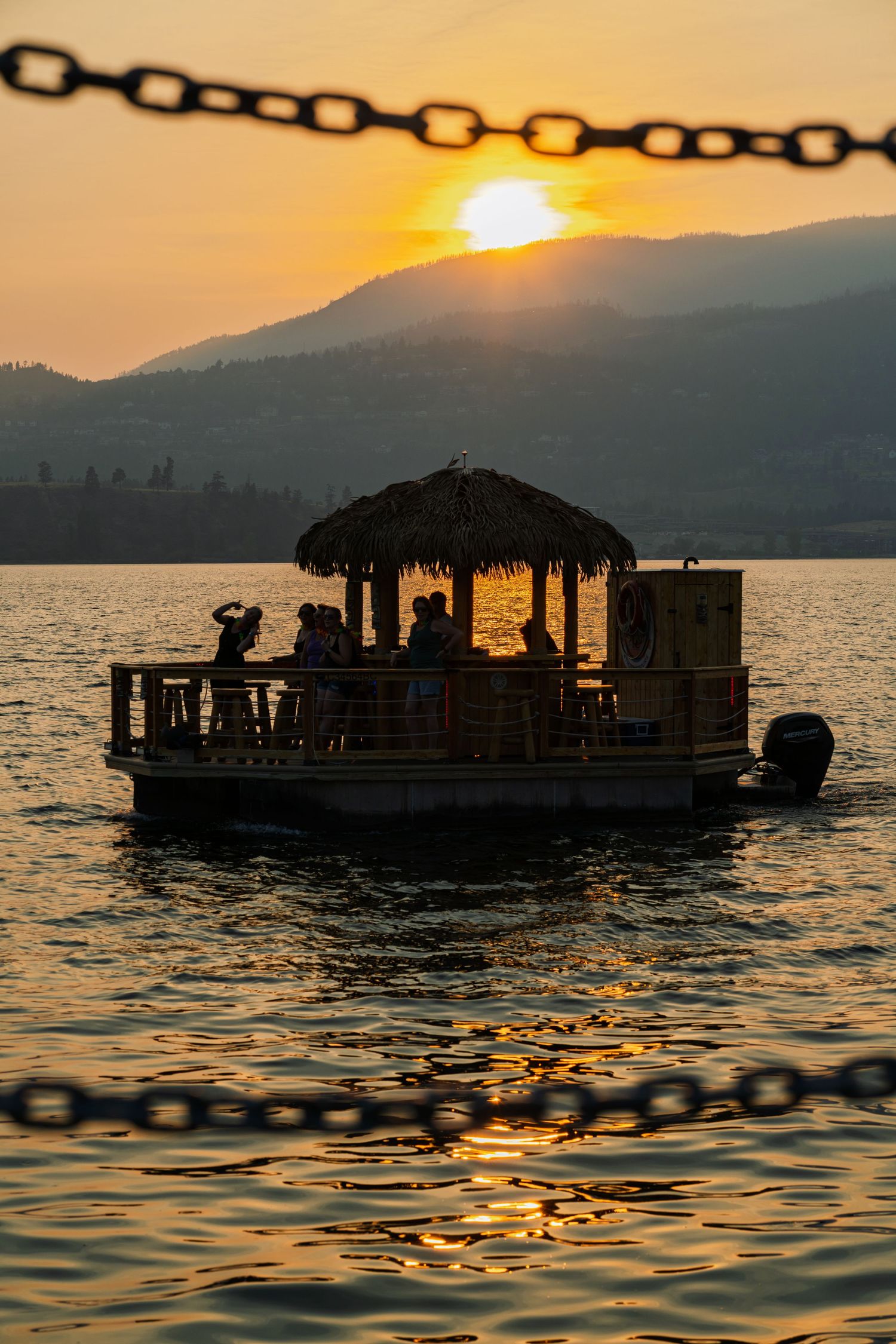 A thatched-roof pontoon boat silhouetted against a golden sunset over mountains and lake.