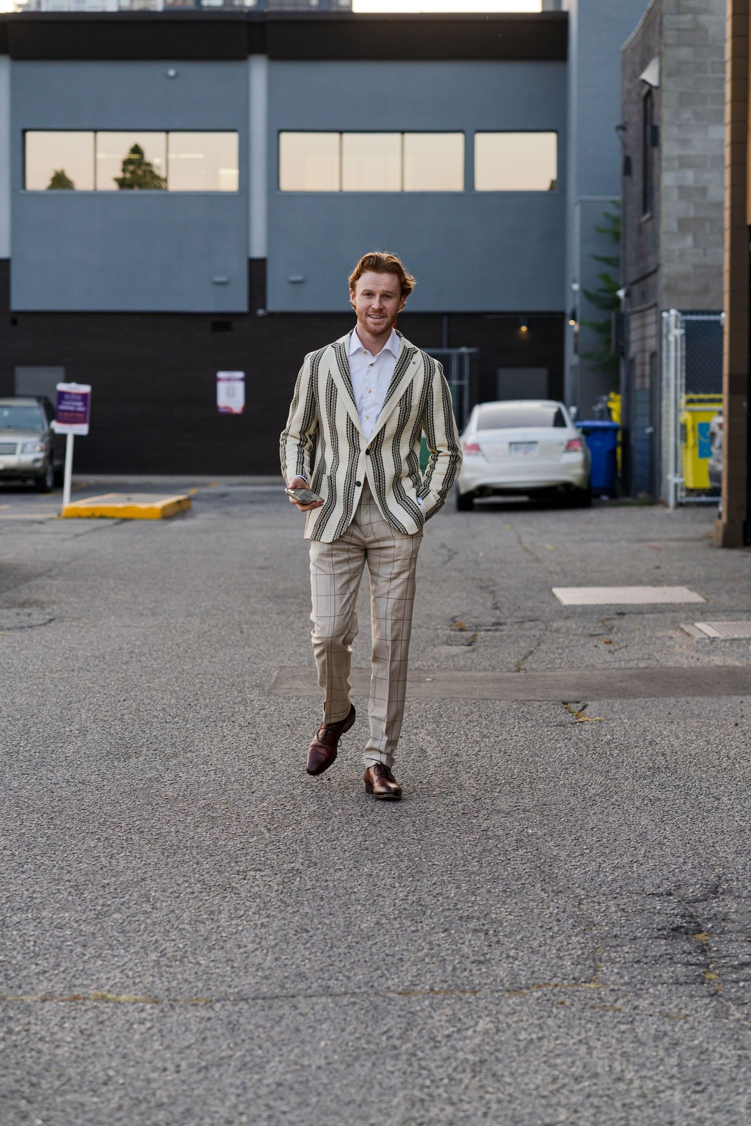 Person wearing striped blazer and light pants walks confidently through urban parking area.