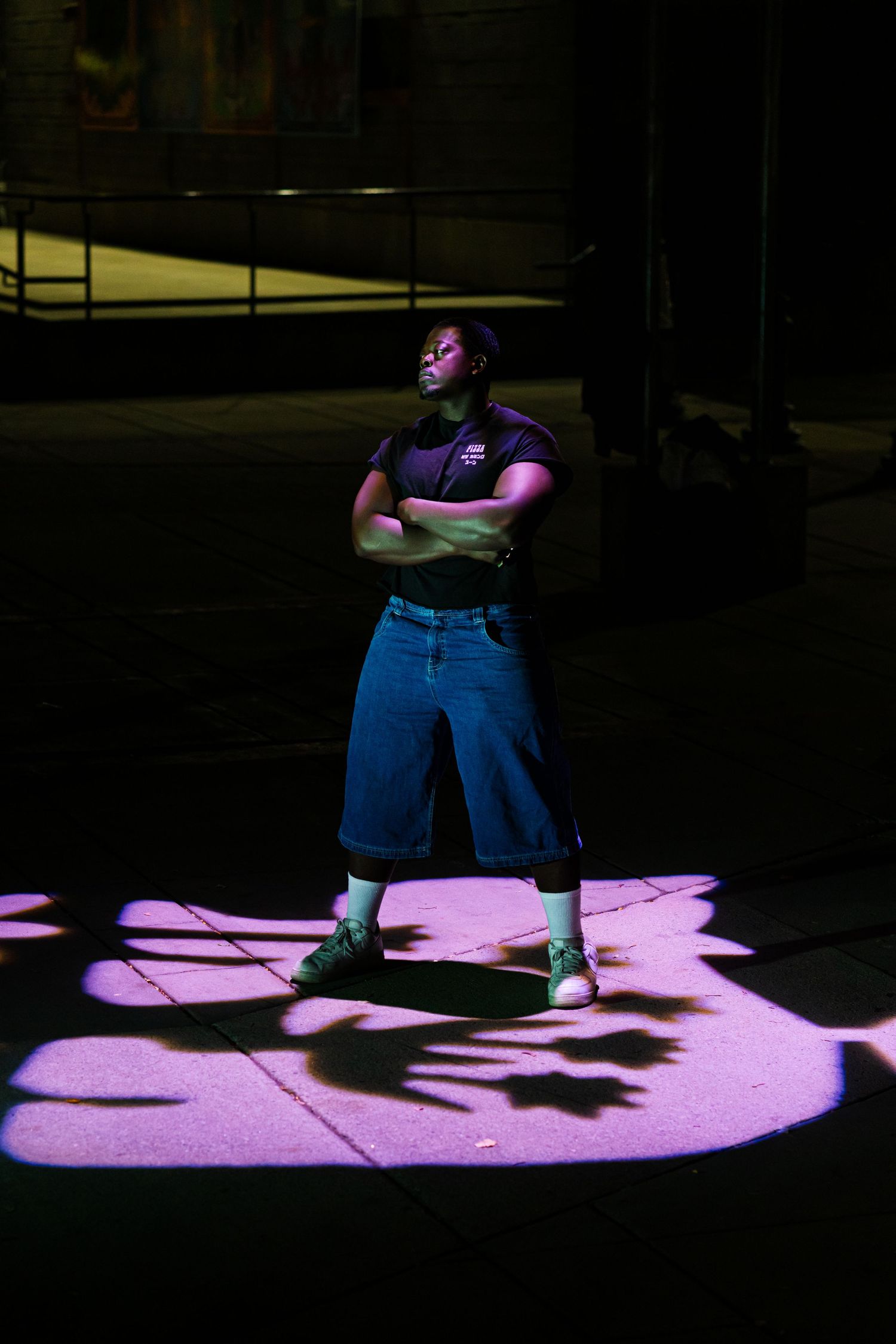 Person standing in dramatic purple lighting on dark stage with crossed arms and casual outfit.