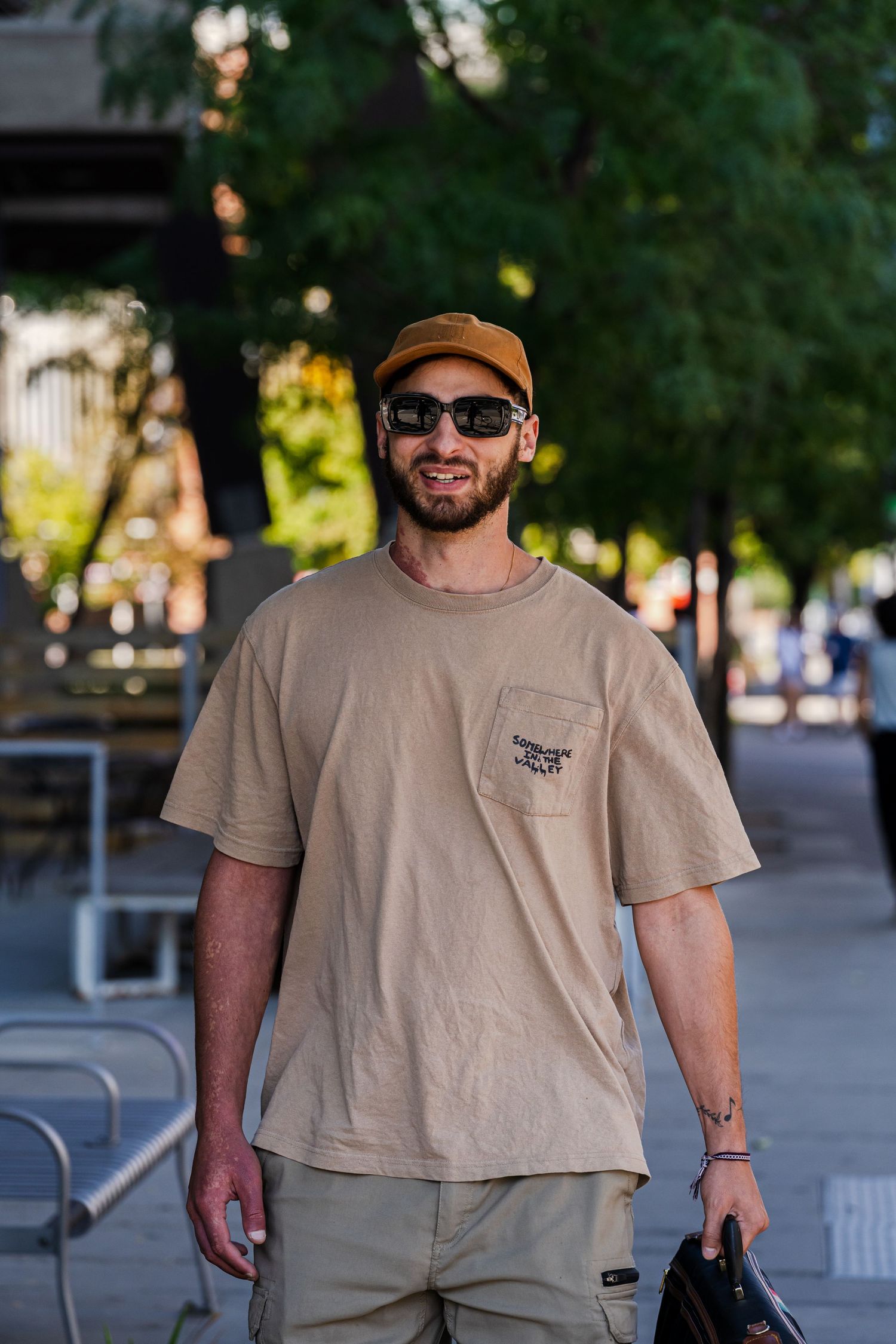 Person walking outdoors wearing tan t-shirt, cap and sunglasses on sunny day.