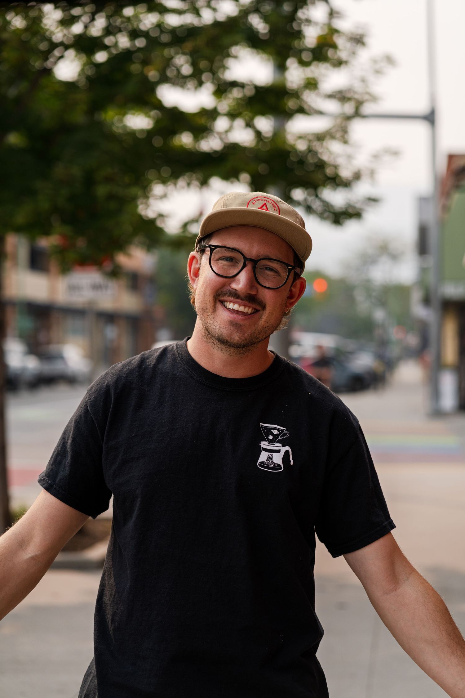 Person in black t-shirt and cap smiles while standing on a city street.
