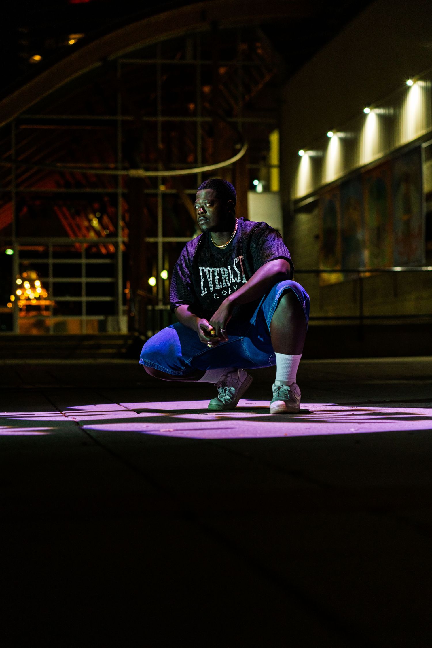 Person in dark clothing crouches on illuminated stage with architectural lighting in background.
