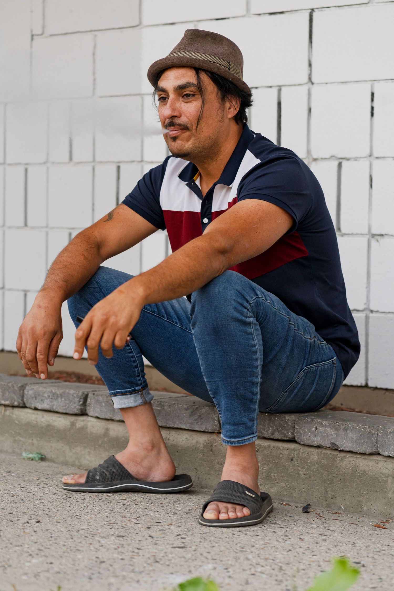 A person in casual attire with a brown fedora sits on a concrete step against a white wall.