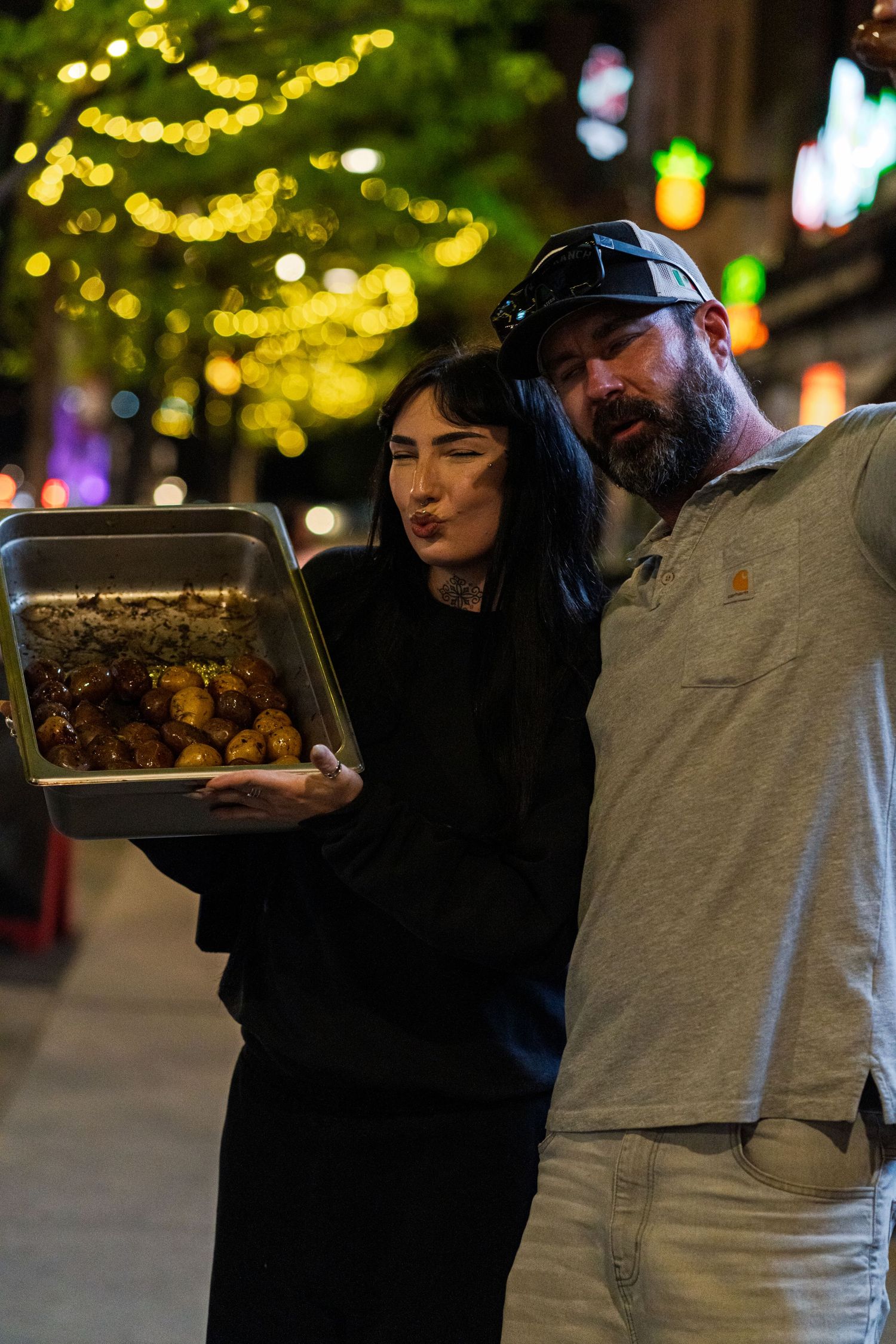 People carry food trays at night under decorative yellow string lights.