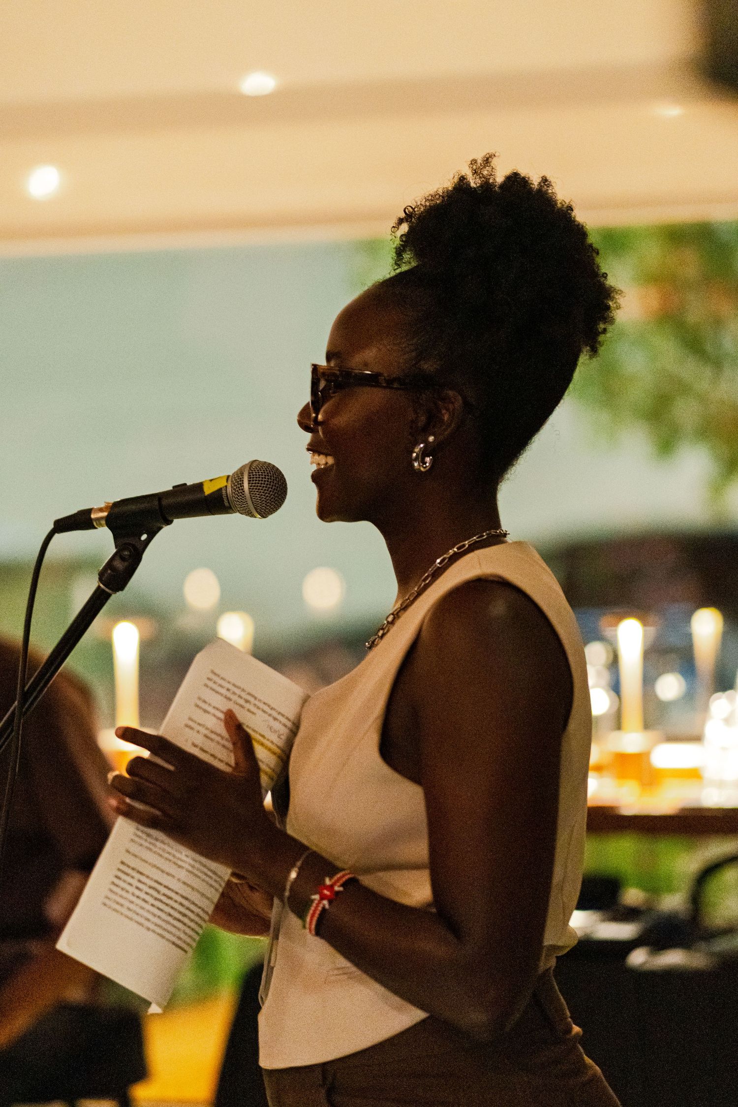 Speaker in white dress holding papers at microphone with candlelit backdrop.