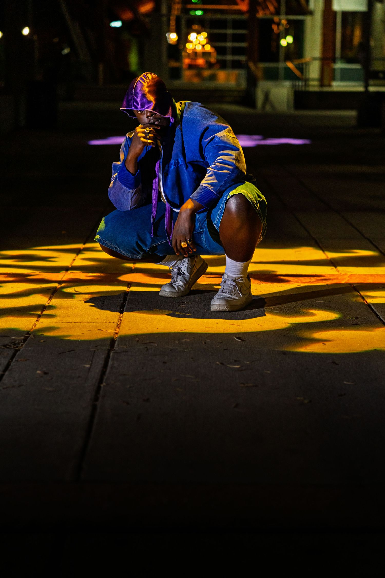 Person in blue streetwear outfit crouching in dramatic nighttime lighting with yellow spotlights on dark pavement.