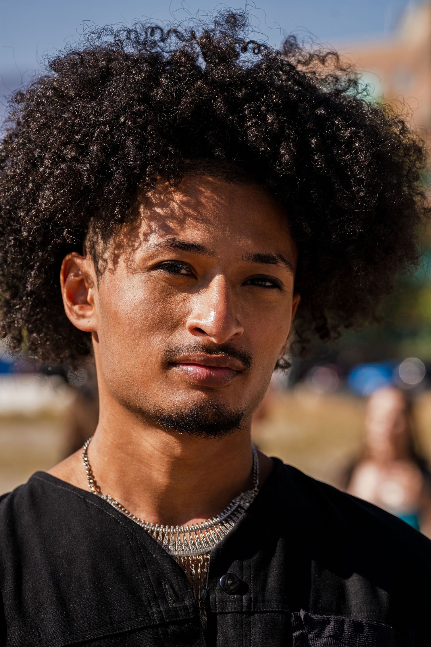 Portrait of person with natural curly afro hairstyle wearing black shirt and silver necklace outdoors.