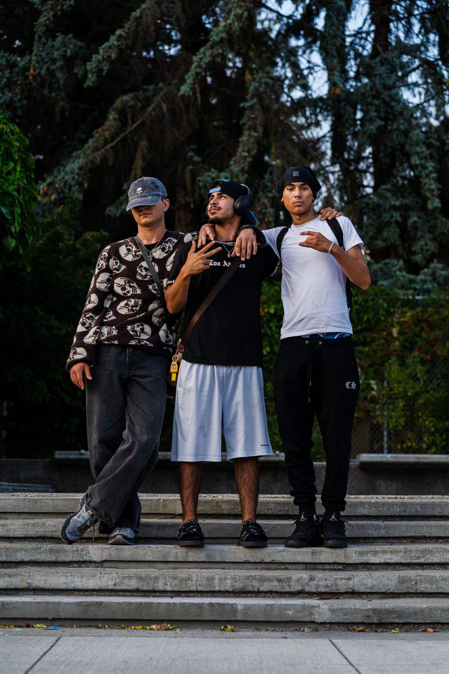 Three friends posing casually on concrete steps in front of evergreen trees in an urban setting.