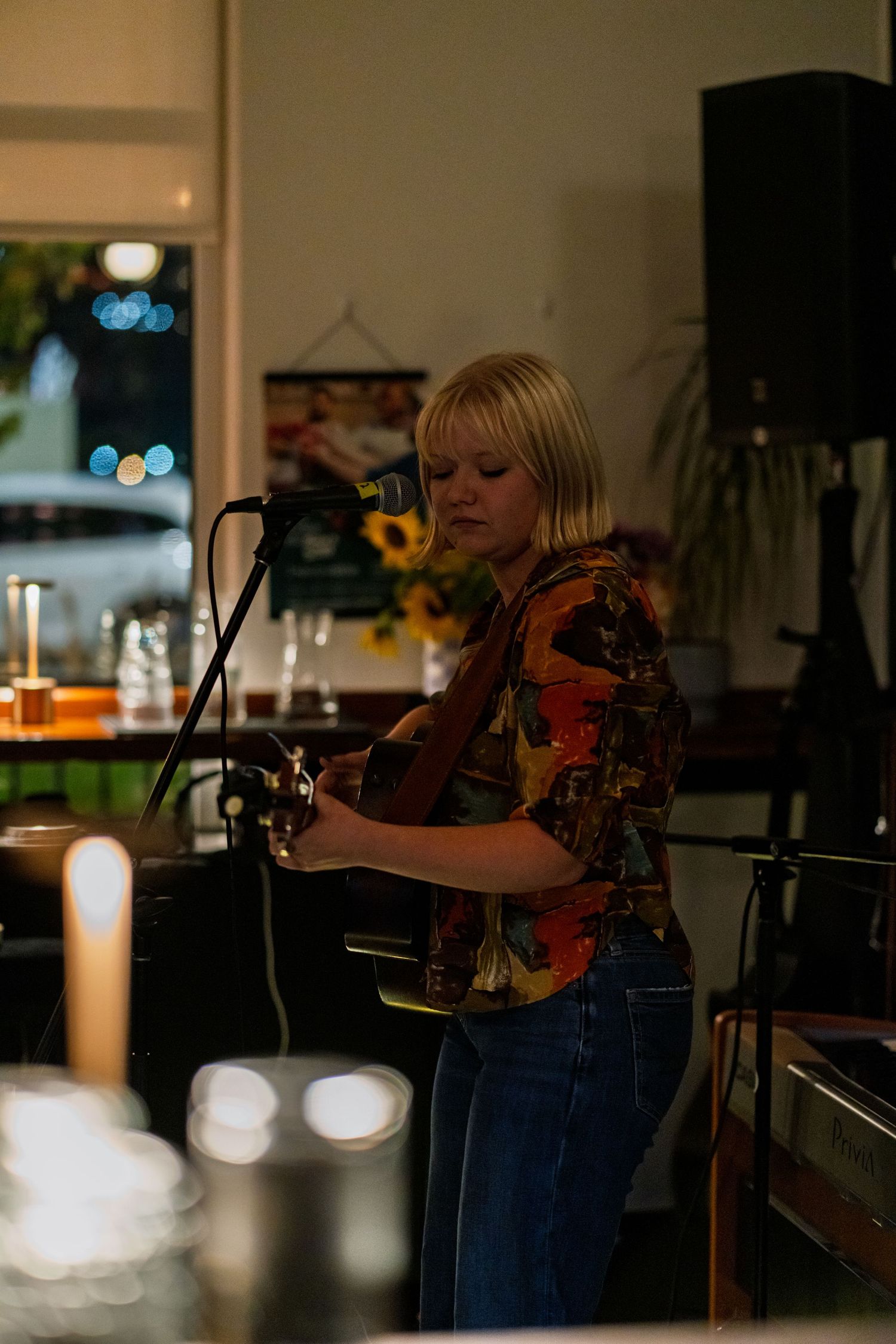 Performer playing acoustic guitar in dimly lit venue with candles and bokeh lighting.