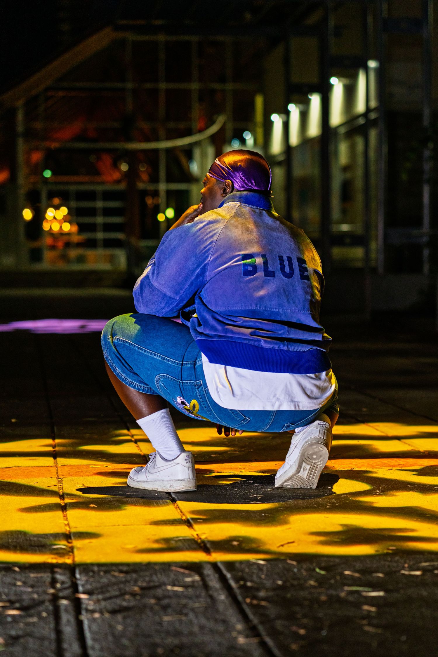 Figure in blue jacket and jeans crouches under urban night lighting with yellow spots on ground.