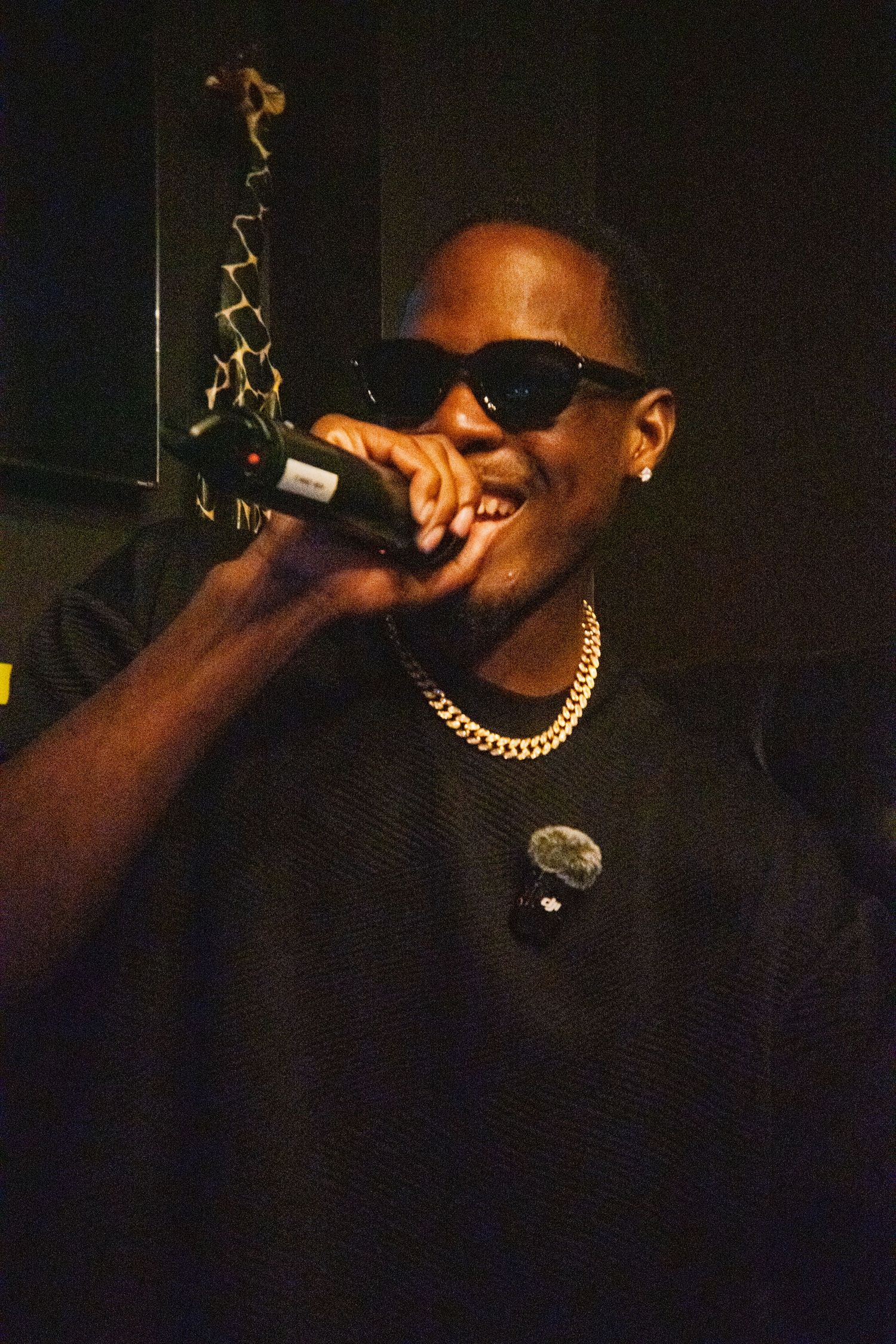 Performer in dark clothing with gold chain performing on stage in low light conditions.