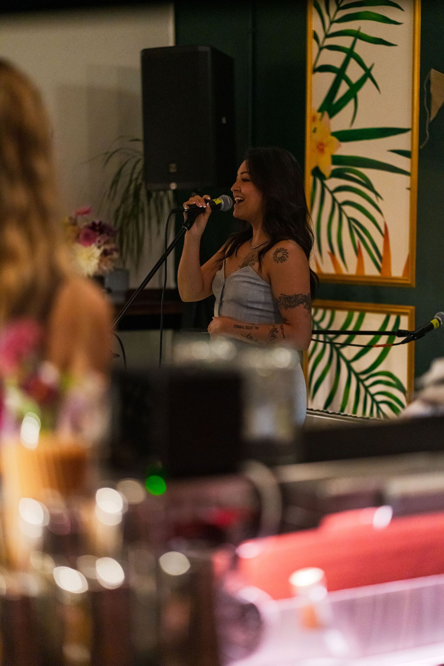Vocalist singing in front of tropical leaf wall art in moody lighting.