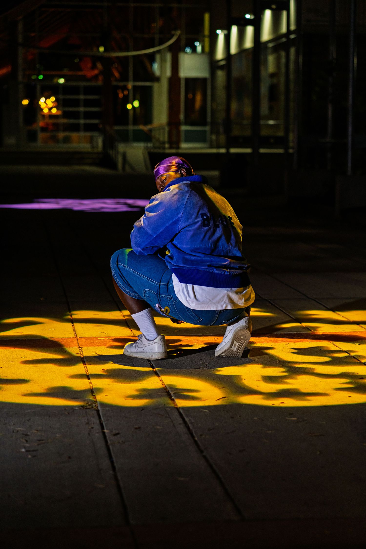 Figure squats on dark stage illuminated by vibrant yellow and purple lighting effects.