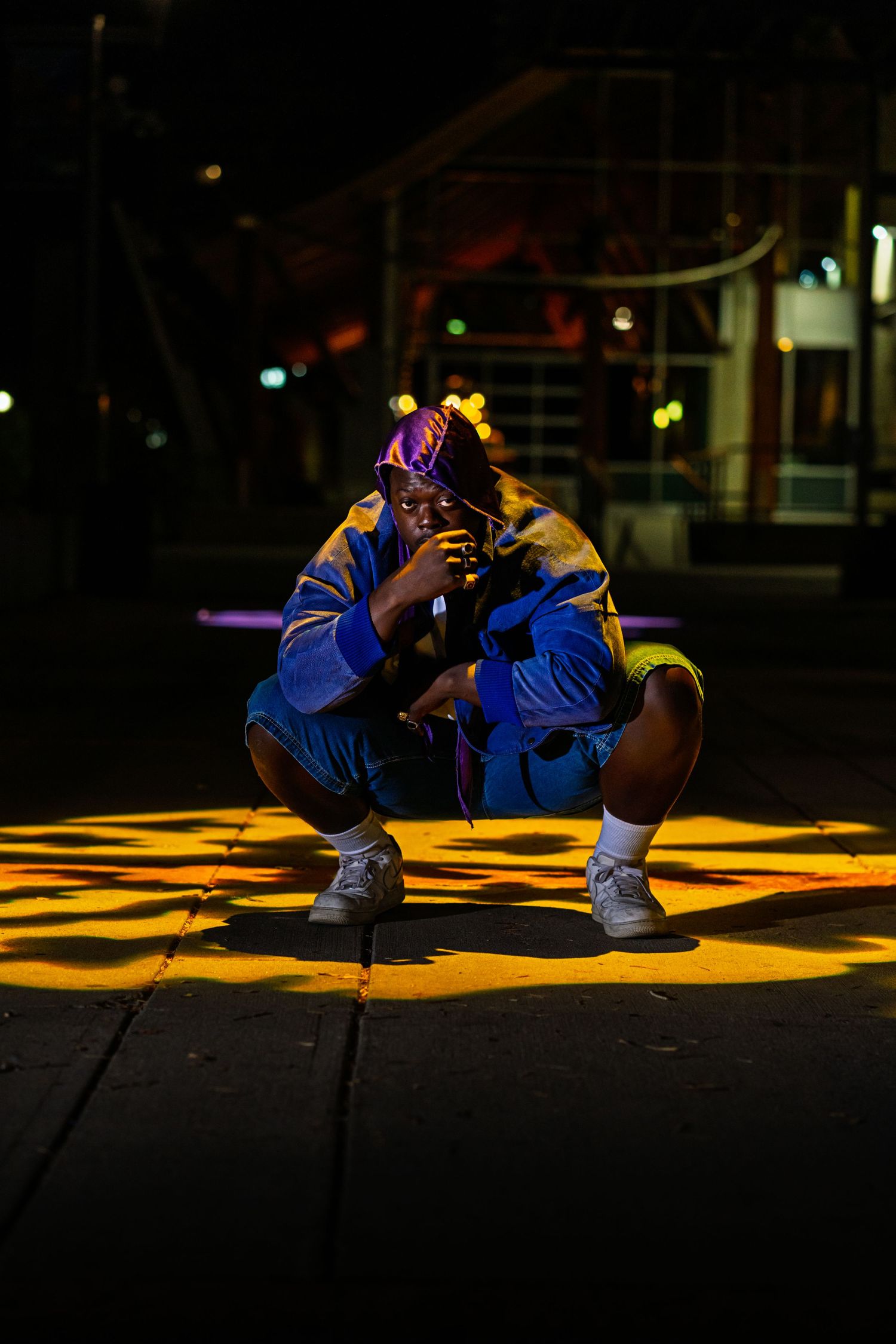 Figure in blue athletic wear crouches in dramatic lighting with yellow spotlights on dark urban ground.