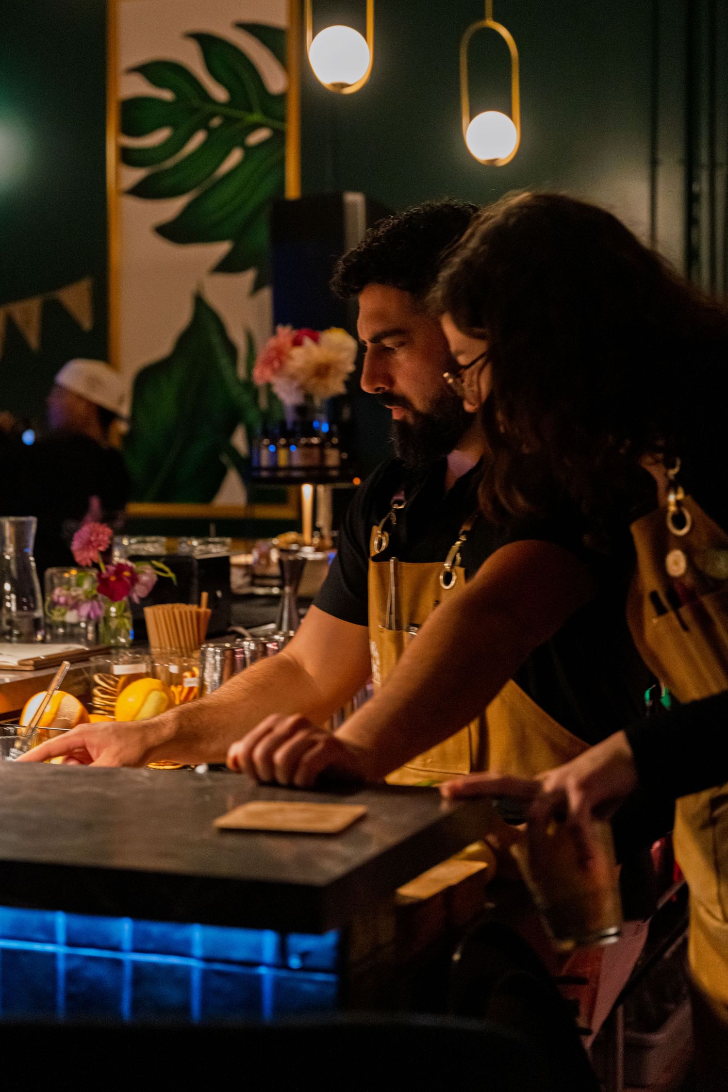 People gather at dimly lit tropical-themed bar with pendant lights and monstera leaf decor.