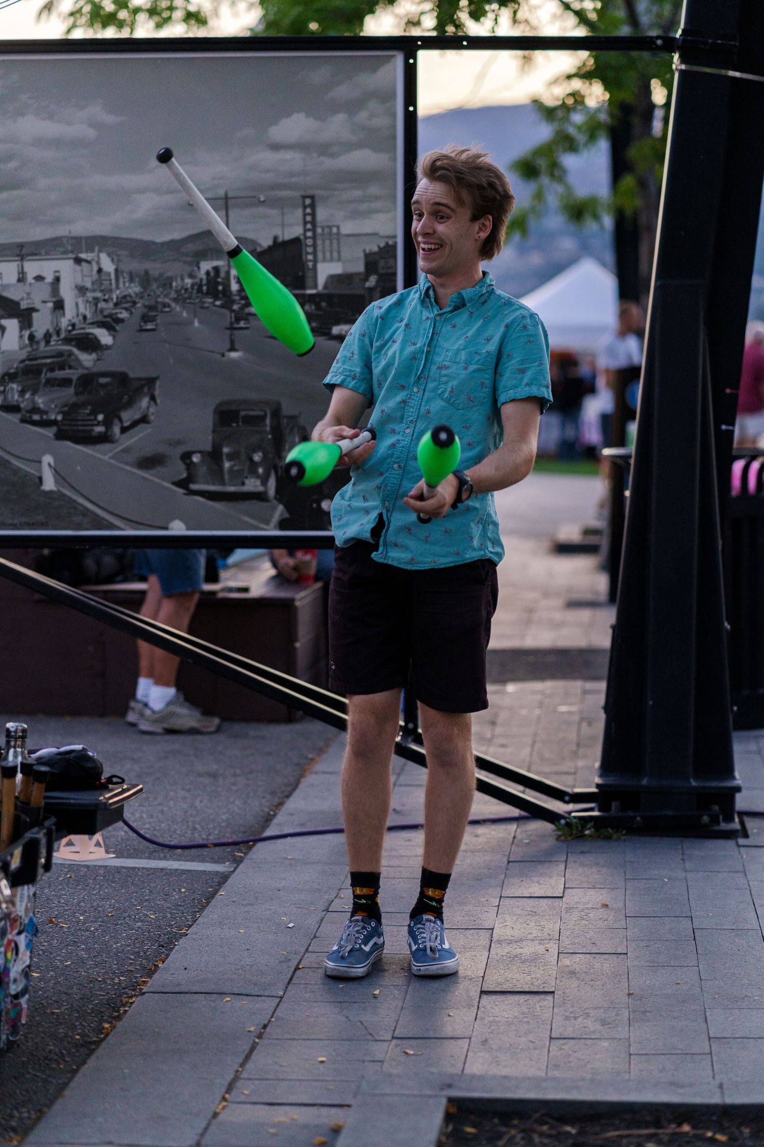 Performer juggling green clubs on street with black and white projection screen in background.