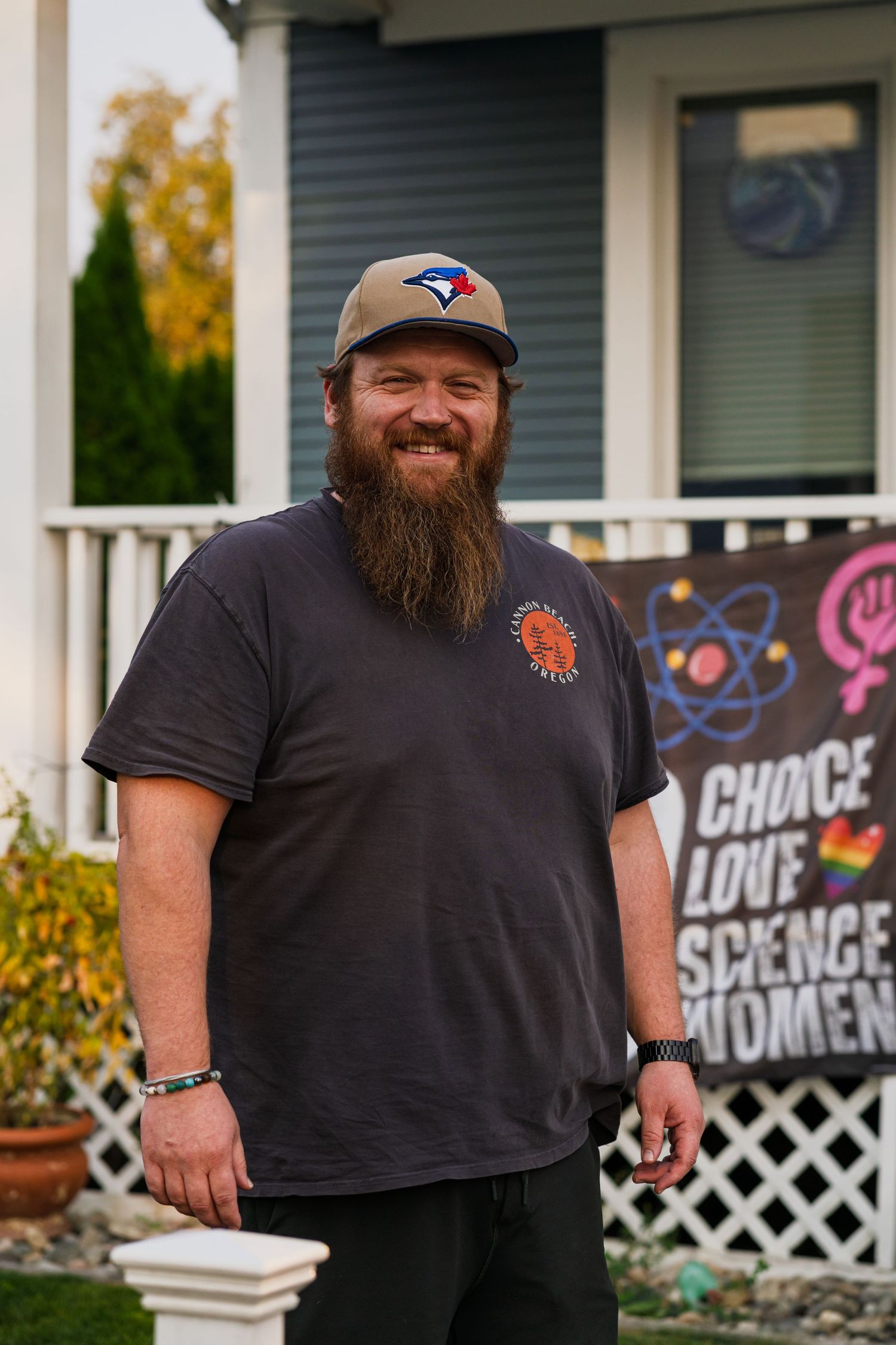 Bearded man in black t-shirt and baseball cap laughing joyfully in front of a house with white lattice fence.