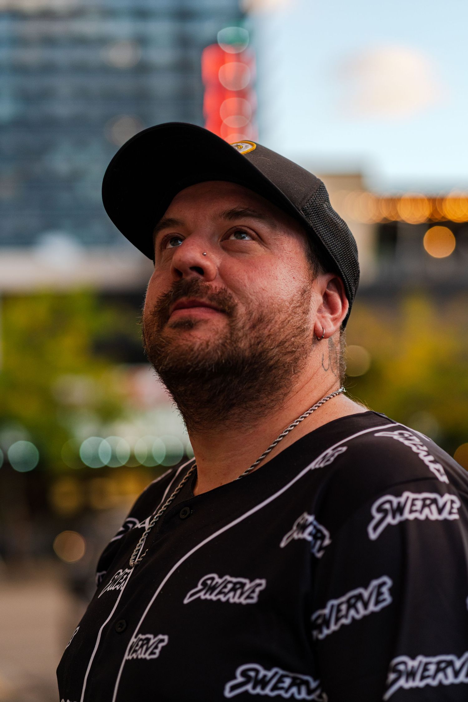 A person wearing a black baseball cap and patterned shirt looks up at the sky with city lights in the background.