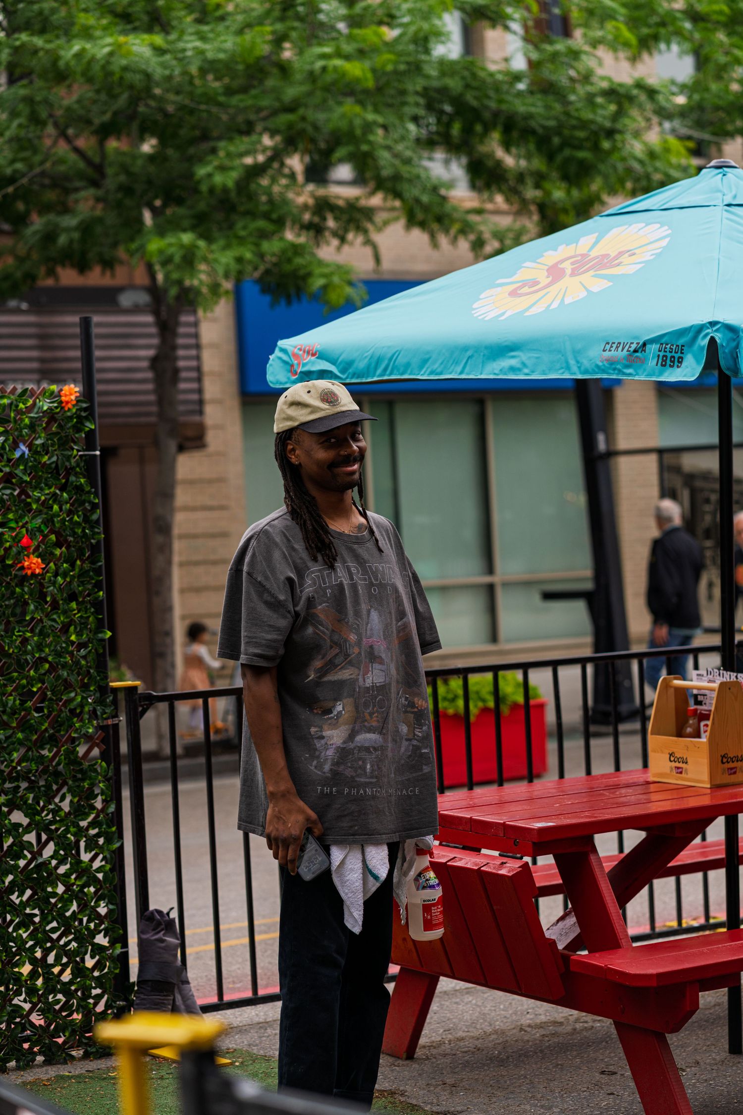 Person stands near a red picnic table under blue umbrellas in an outdoor setting.
