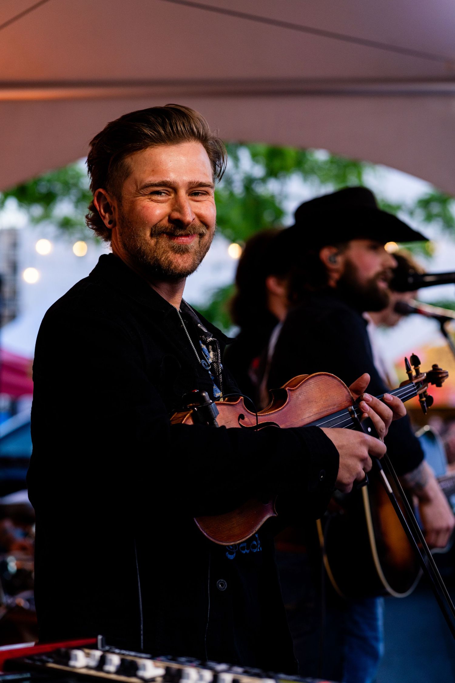 Musician smiling while holding violin under tent at outdoor performance.
