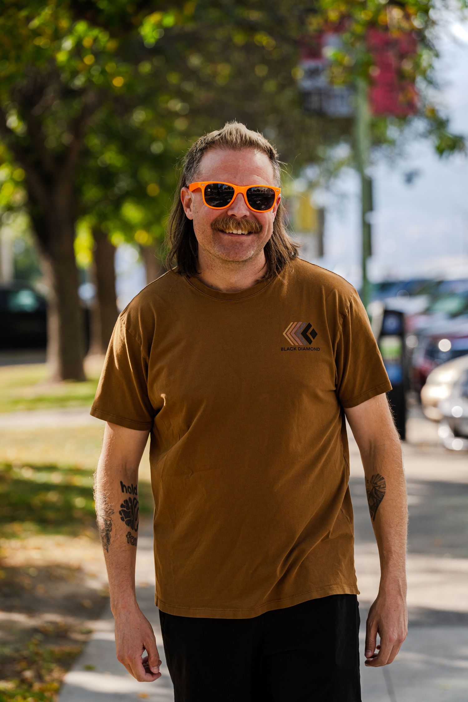 Person in brown t-shirt and sunglasses poses outdoors on sunny day.