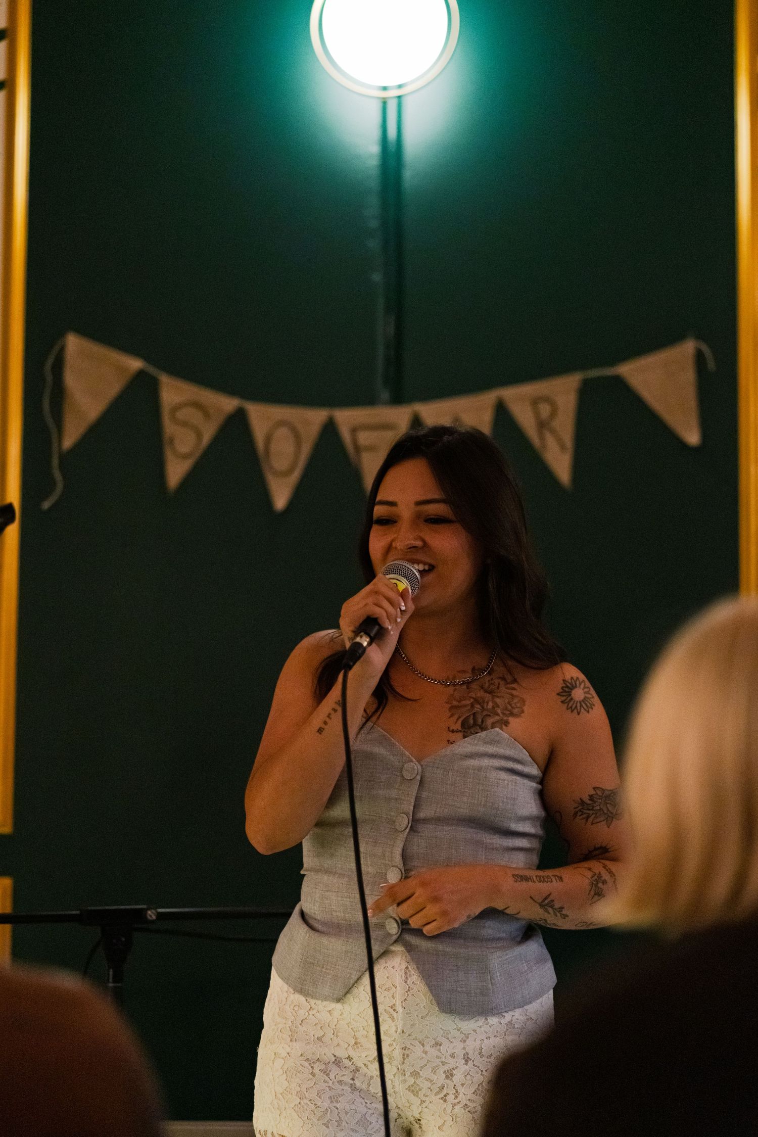 A performer in a white dress sings passionately into a microphone at a dimly lit venue with string lights and bunting.