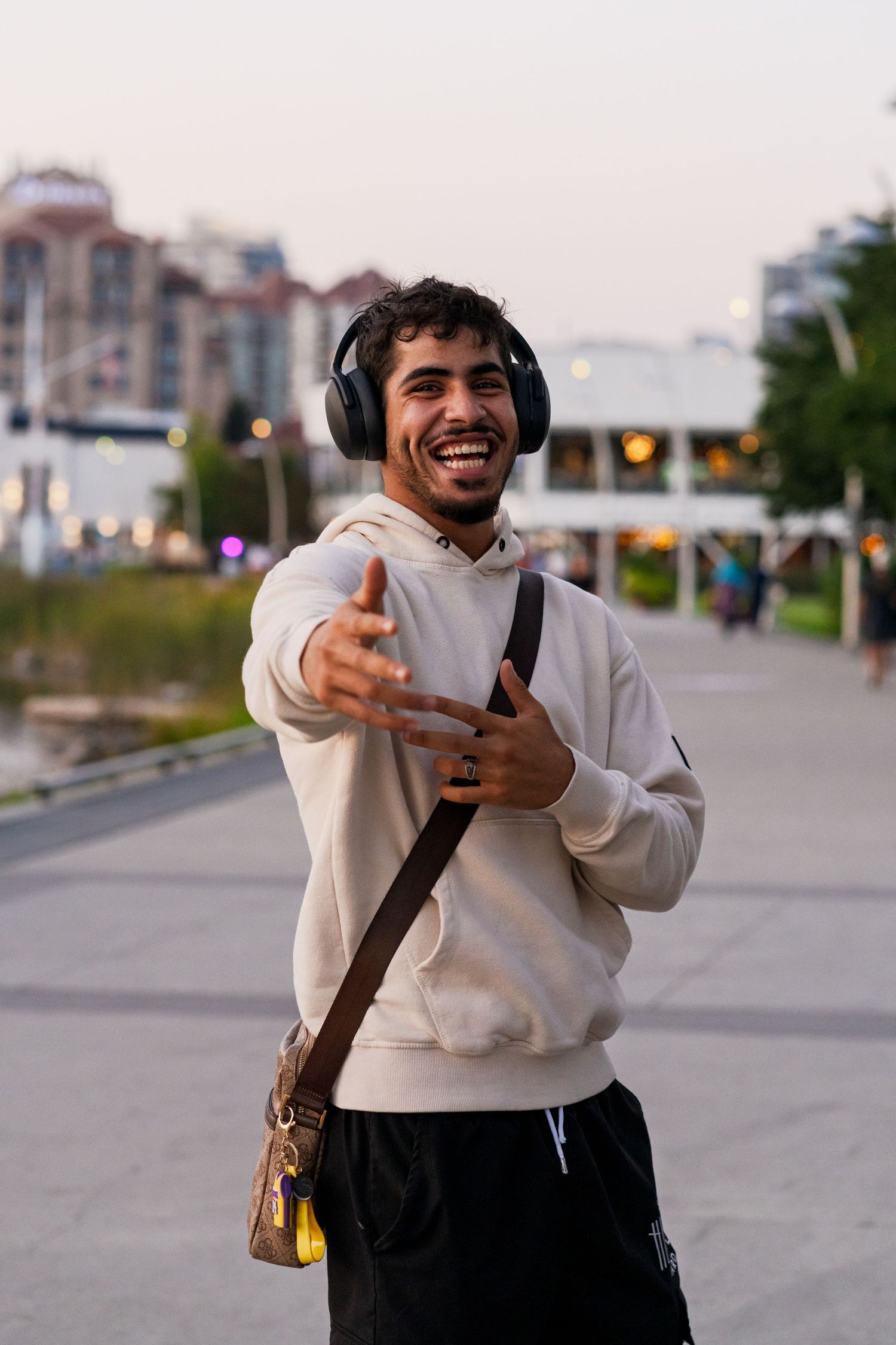 Person wearing headphones and cream hoodie smiling and gesturing while standing on urban path.