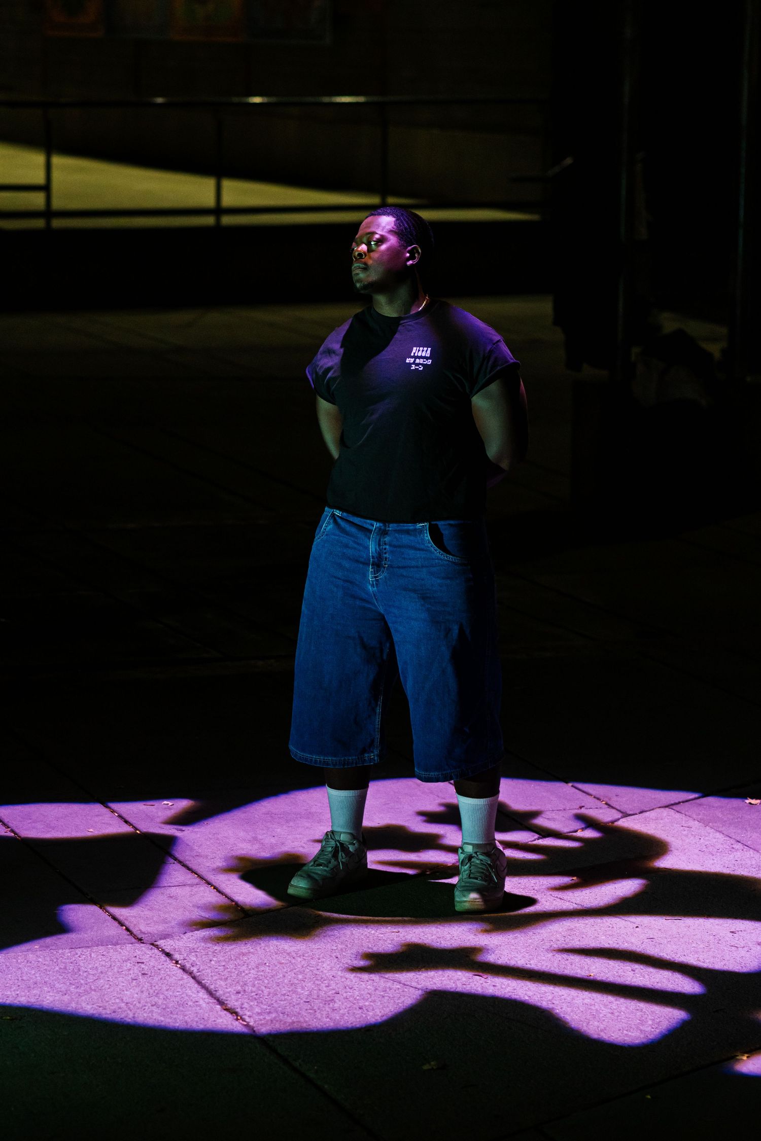 A figure stands in dramatic purple lighting casting shadows on the ground while wearing dark clothing and shorts.