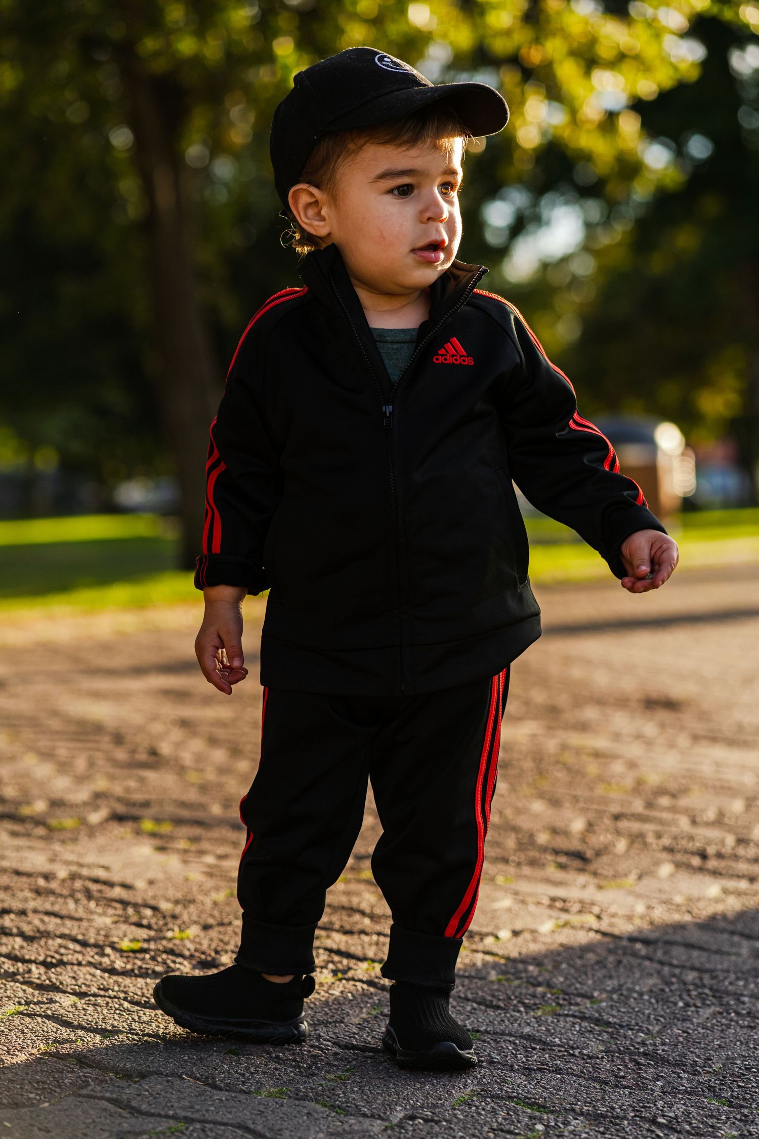 Small child wearing black Adidas tracksuit with red stripes stands outdoors at sunset.