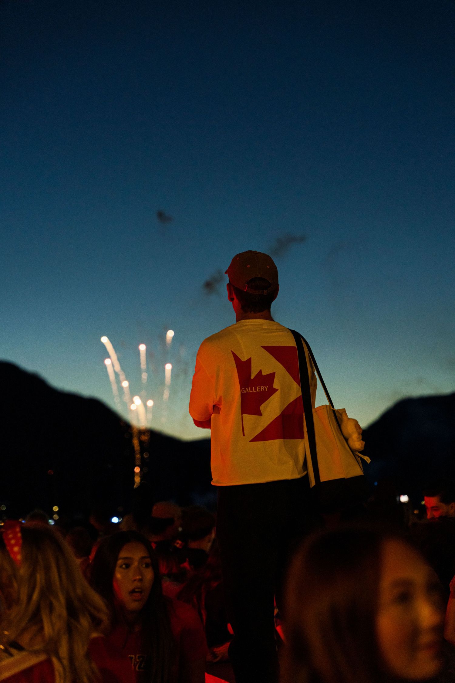 Person in orange maple leaf jersey watching fireworks against a dark blue sky at dusk.