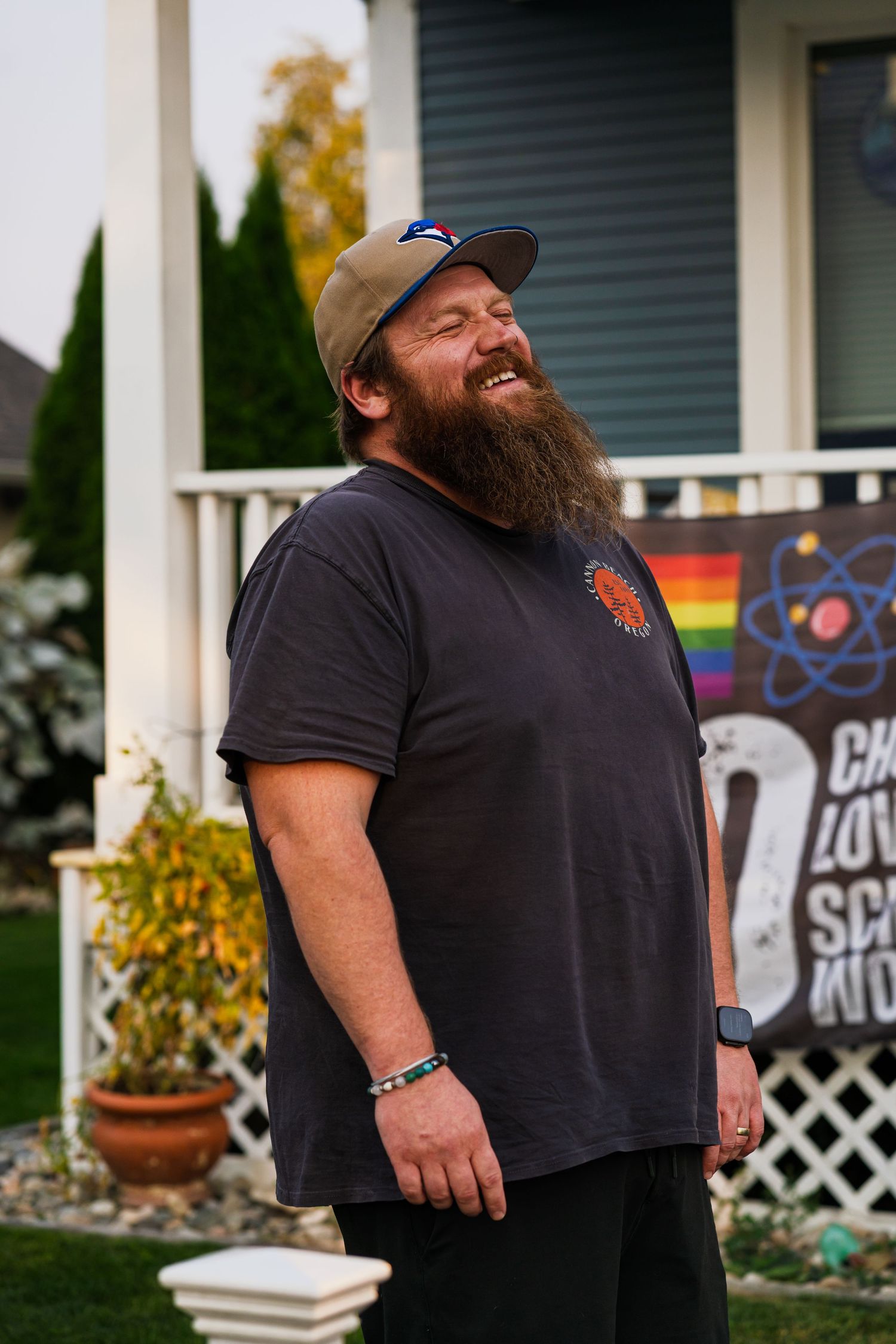 Bearded man in black t-shirt and baseball cap smiling outside a house with white lattice and decorative signs.
