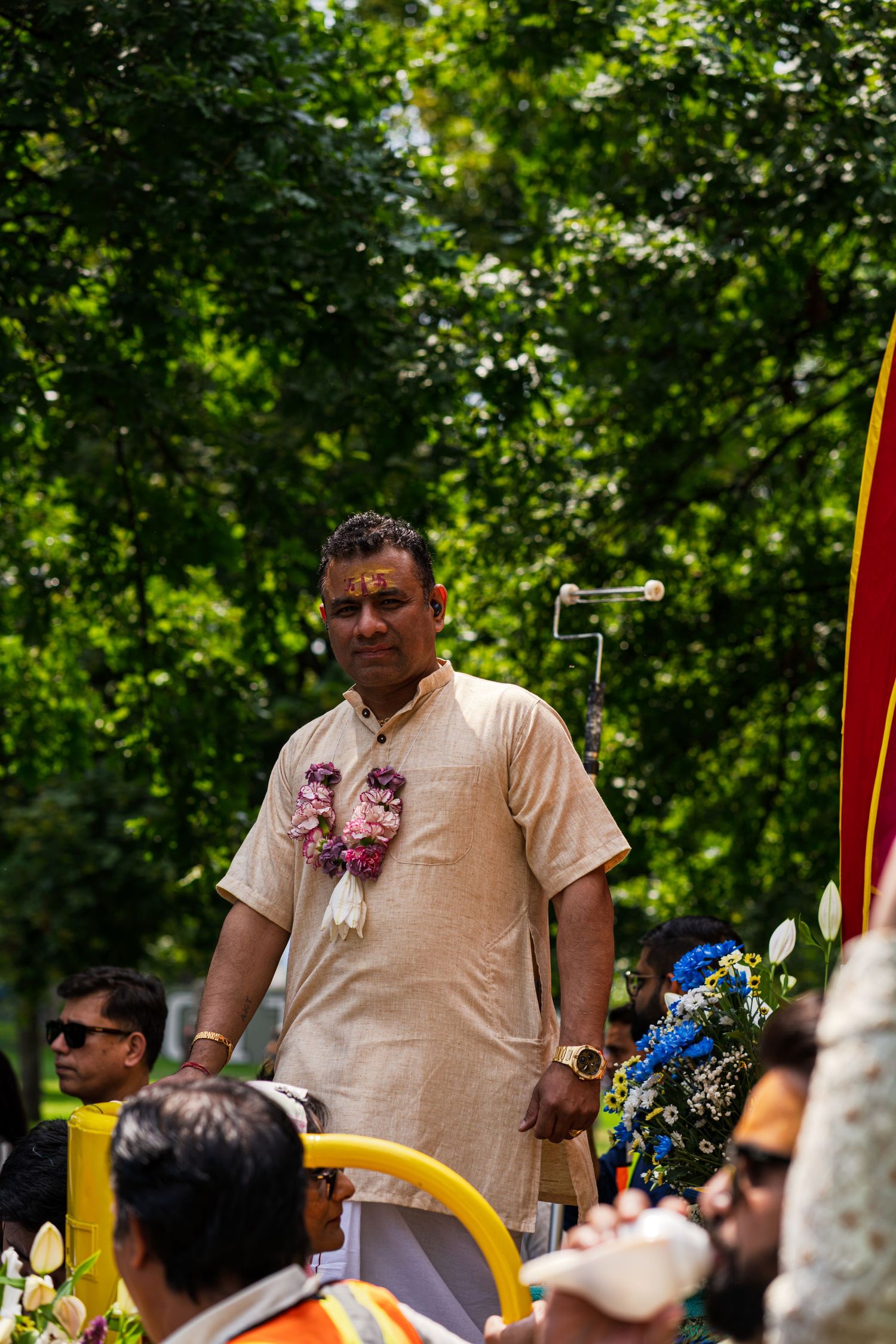 People gather at an outdoor event with trees and flags visible in the background.