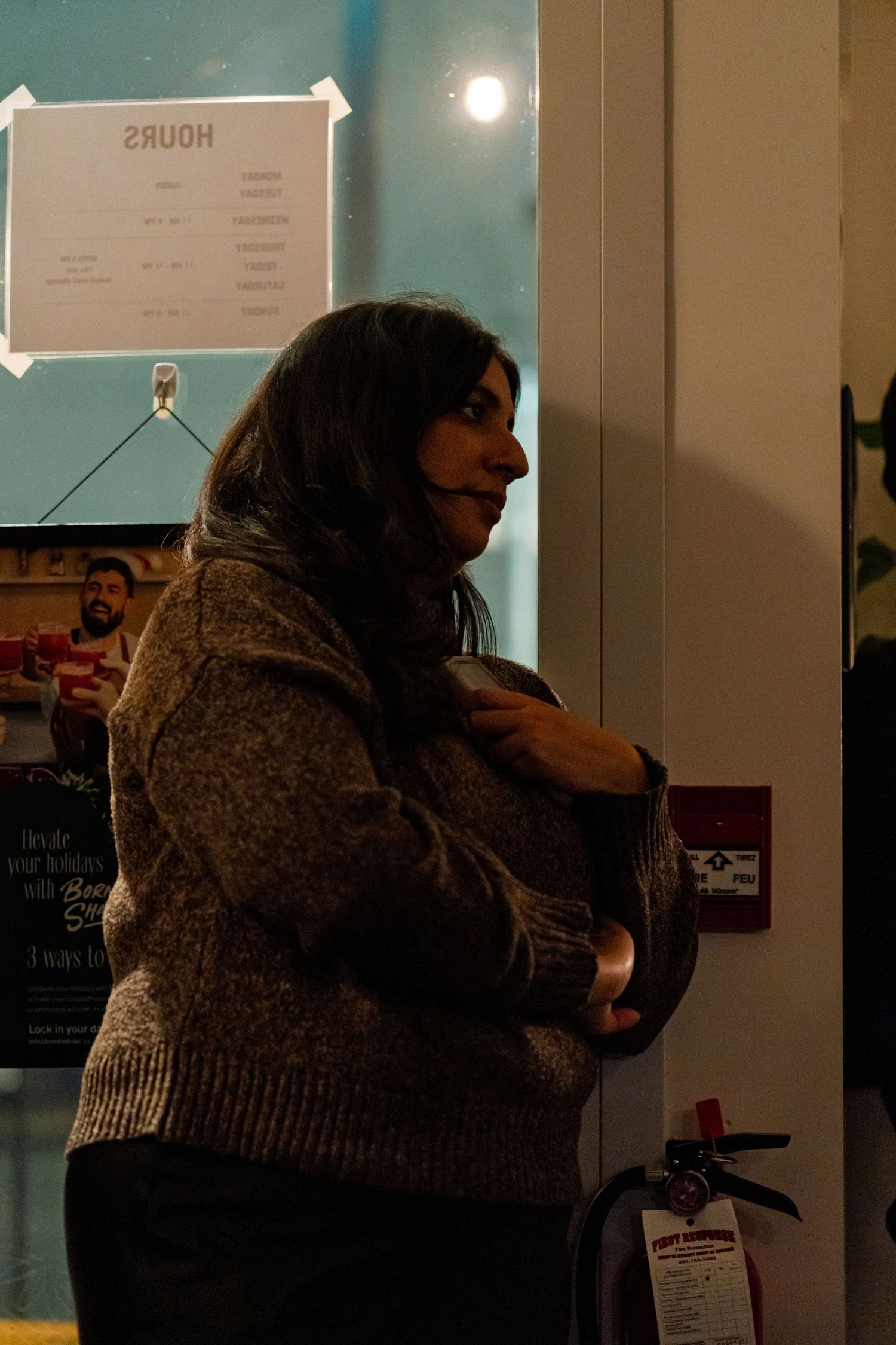 Person in brown sweater standing in dimly lit room next to wall with posted schedule.