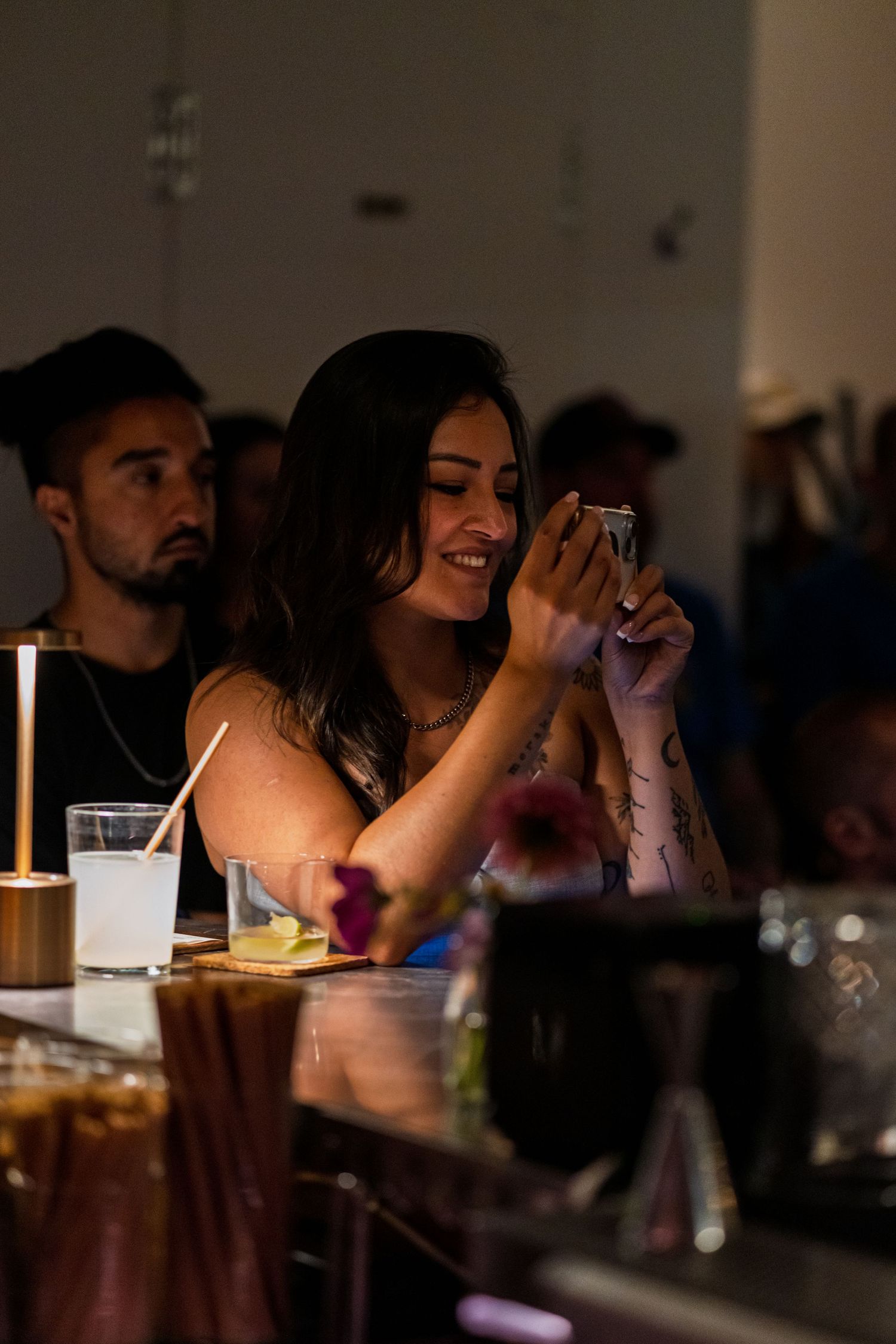Person at dimly lit bar taking photo of cocktail with phone.