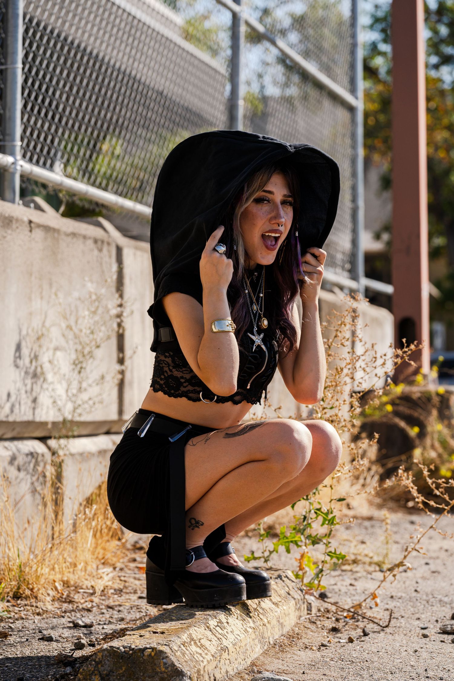 Woman in black outfit and wide-brimmed hat crouches while posing outdoors.