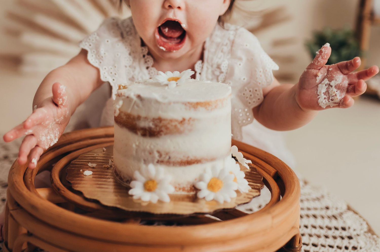 Baby girl in white dress excitedly smashing her first birthday cake decorated with daisy flowers.