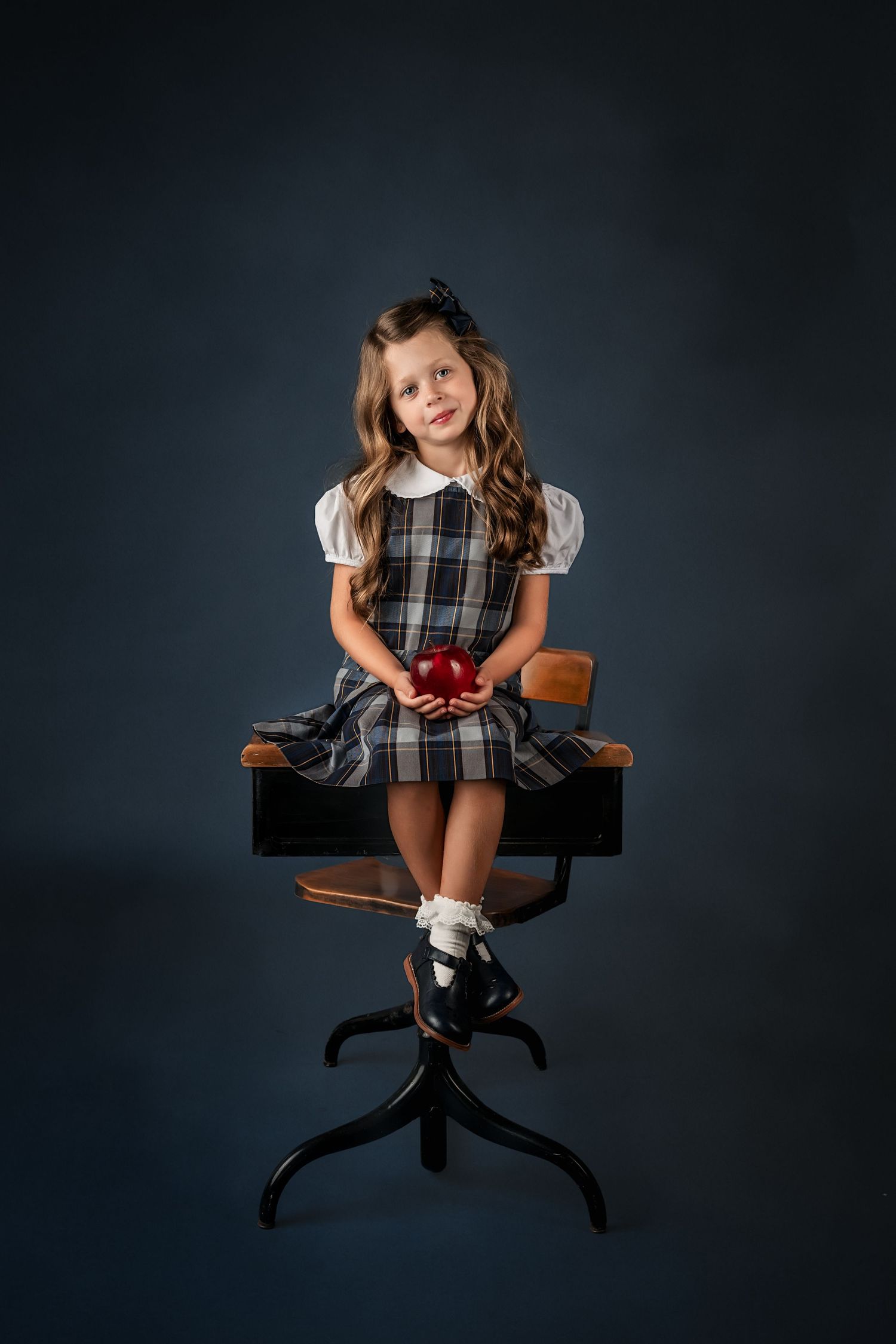 Young girl in plaid school uniform sitting on vintage desk holding red apple, for fine art studio portrait on dark background in Atlanta.