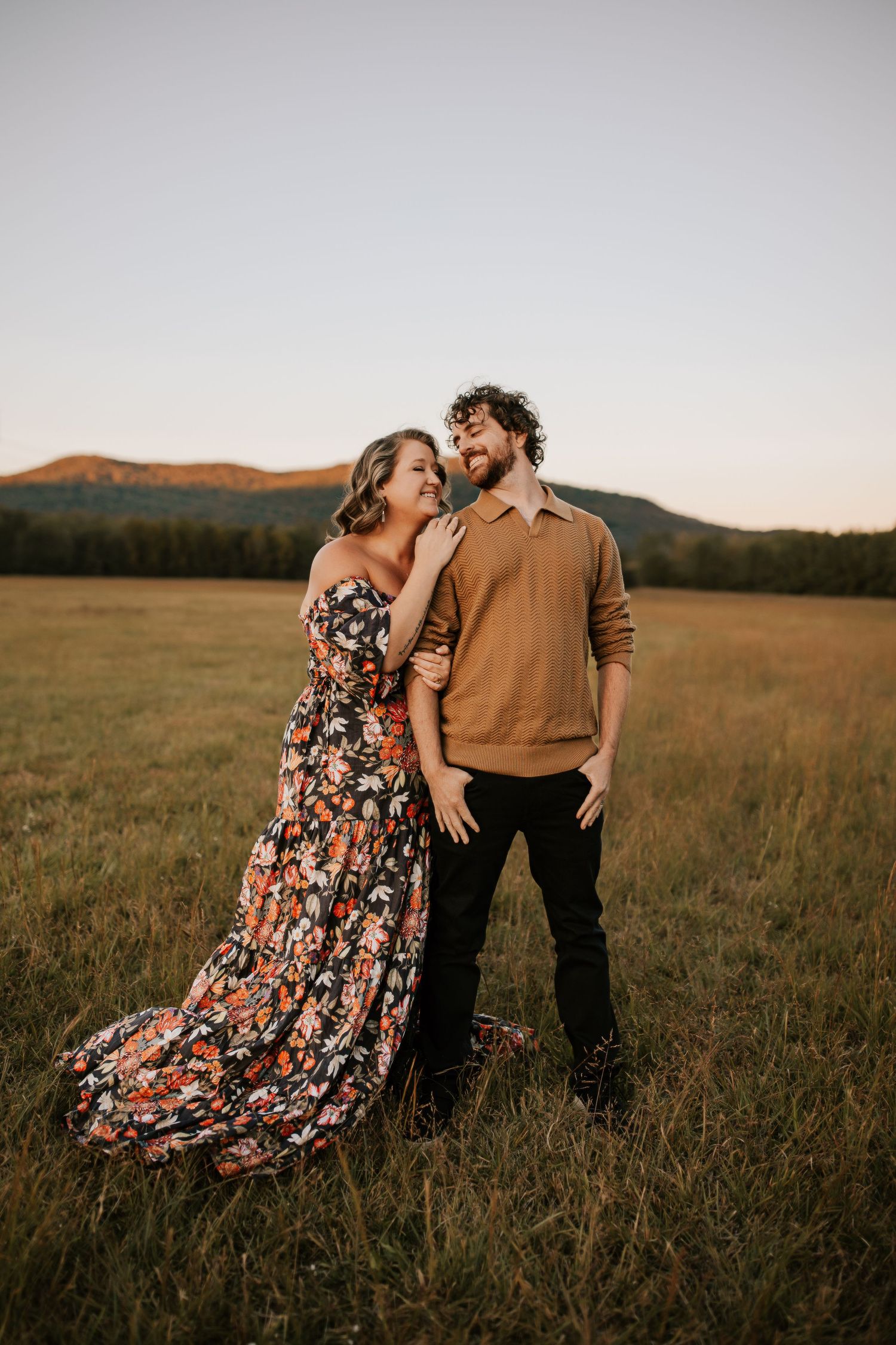A couple embraces in a golden field at sunset wearing a floral dress and brown shirt.