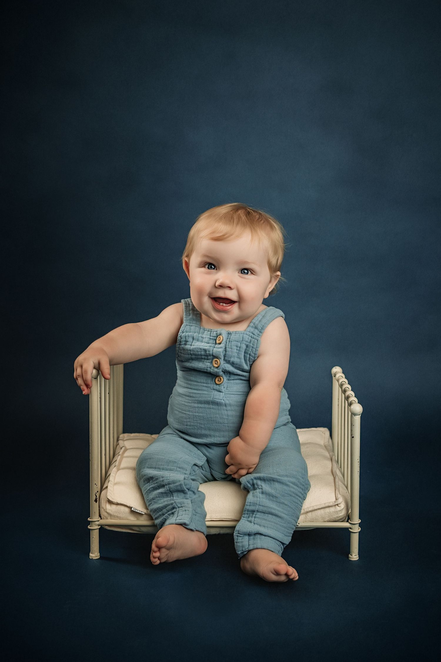 Smiling baby in blue linen romper sitting on a miniature white bed against a dark blue studio background for fine art milestone photos