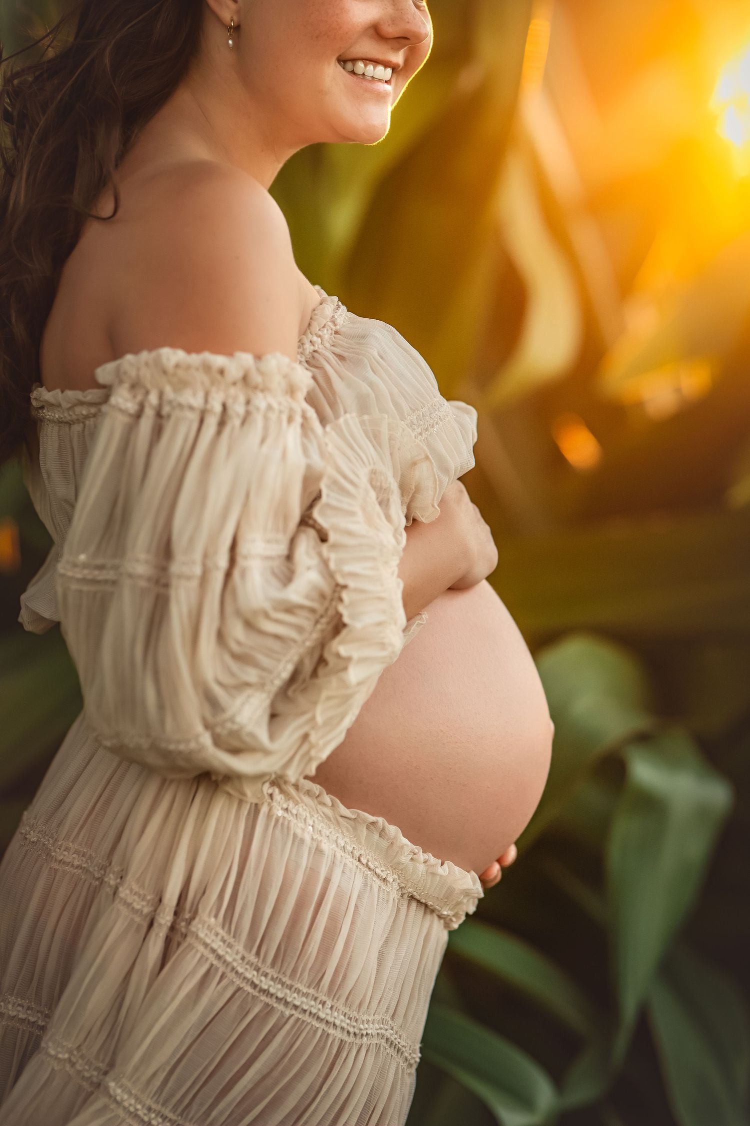 Smiling pregnant woman in off-shoulder cream boho dress cradling bare baby bump outdoors in golden sunset light.