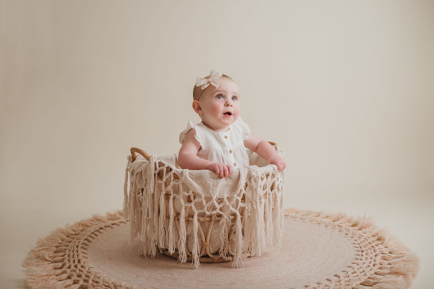 Baby girl in white romper sitting in macrame basket on crochet rug, looking up with bow headband.