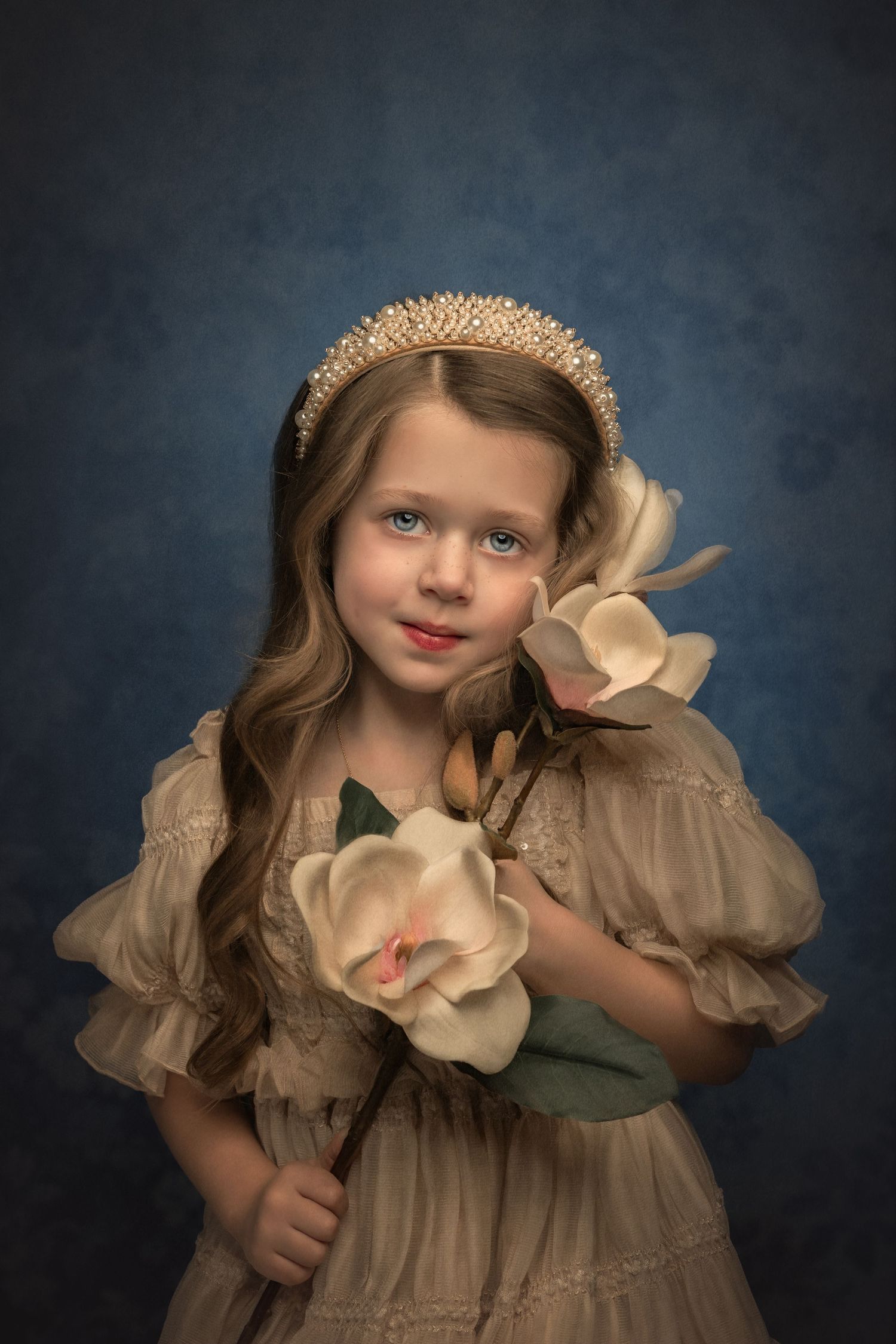 A young girl in a beige dress holds magnolia flowers, wearing a pearl tiara on a blue backdrop.