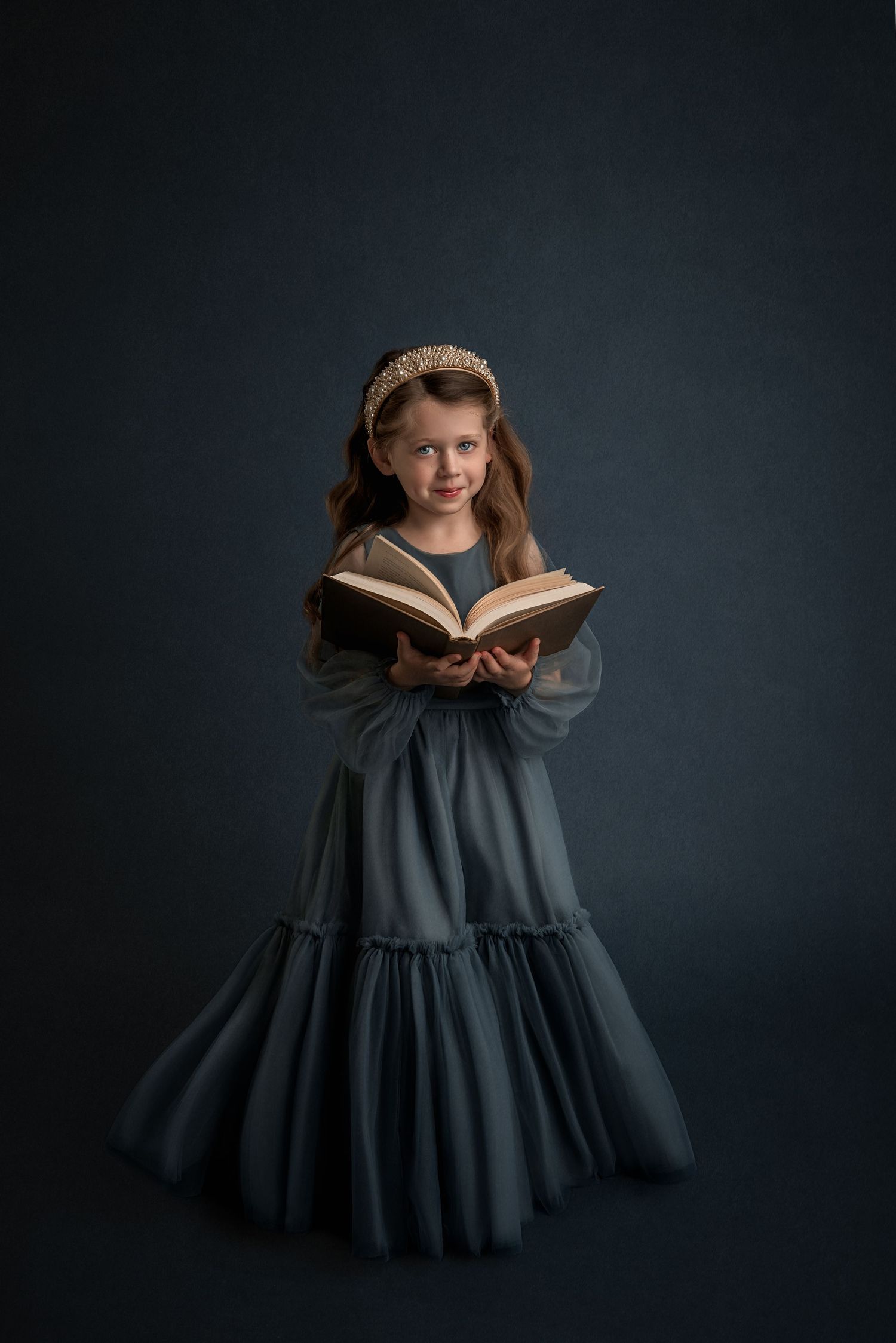 Young girl in teal gown holding open book, wearing pearl headband, posing elegantly against dark blue background.