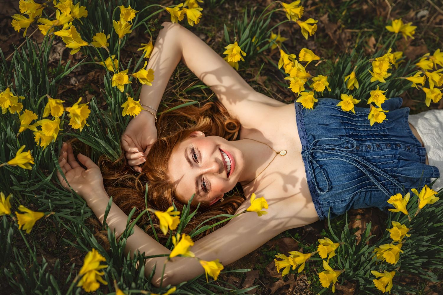 Smiling redhead woman lying among yellow daffodils in a field, wearing a denim top, enjoying a sunny spring day.