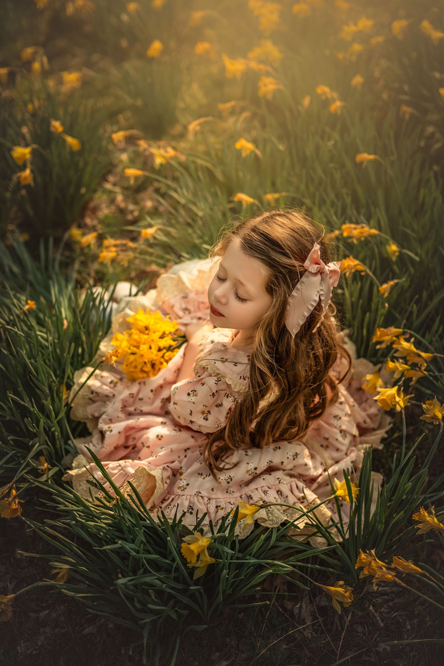 Young girl in floral dress sitting among yellow daffodils, holding flowers, with golden sunlight and a bow in her hair.