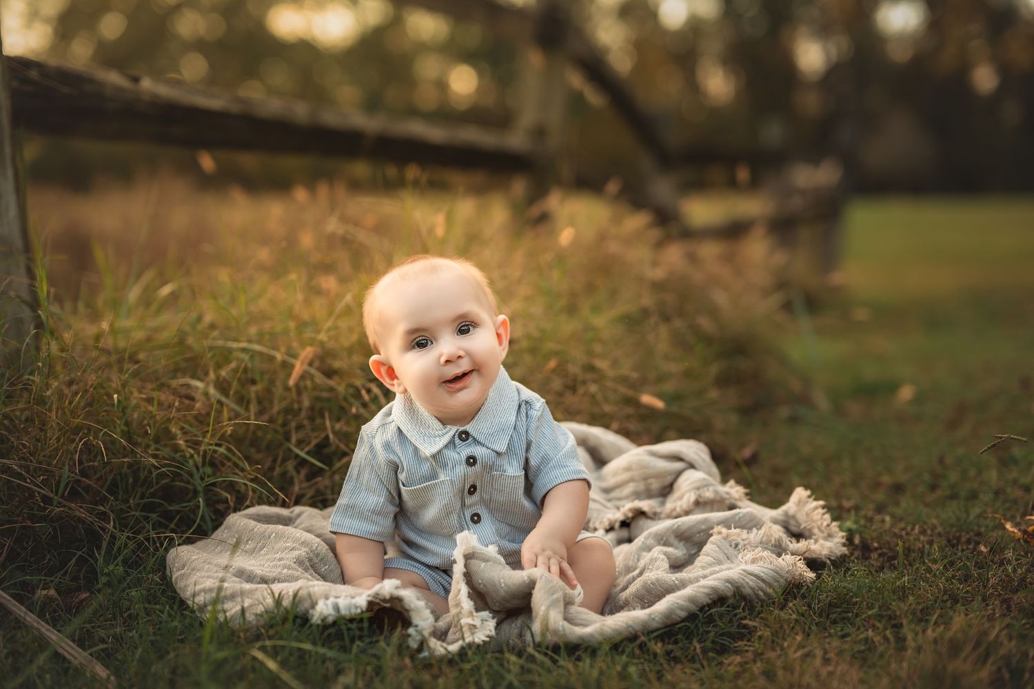 A happy baby sitting on a blanket in a grassy outdoor setting during golden hour.
